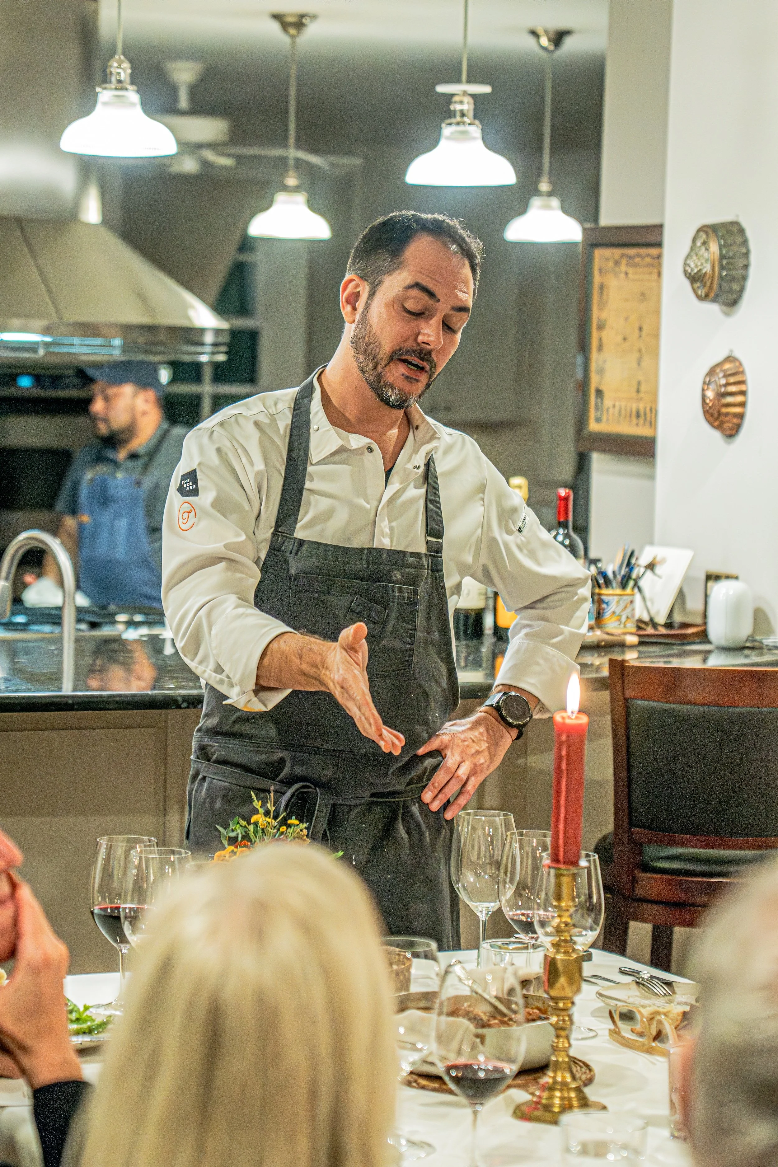 A chef in a white uniform and black apron demonstrates cooking at a dinner event. People are seated at a table with wine glasses and a red candle, and the setting has a cozy, warm ambiance with decorative wall plates.