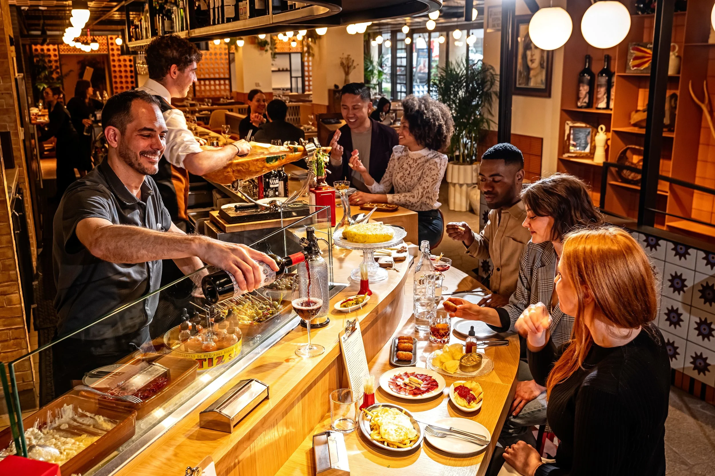 A lively restaurant scene with several people seated at a bar and tables, enjoying food and drinks. The bartender is pouring wine, and the atmosphere is warm and inviting with decorative lighting and artwork on the walls.