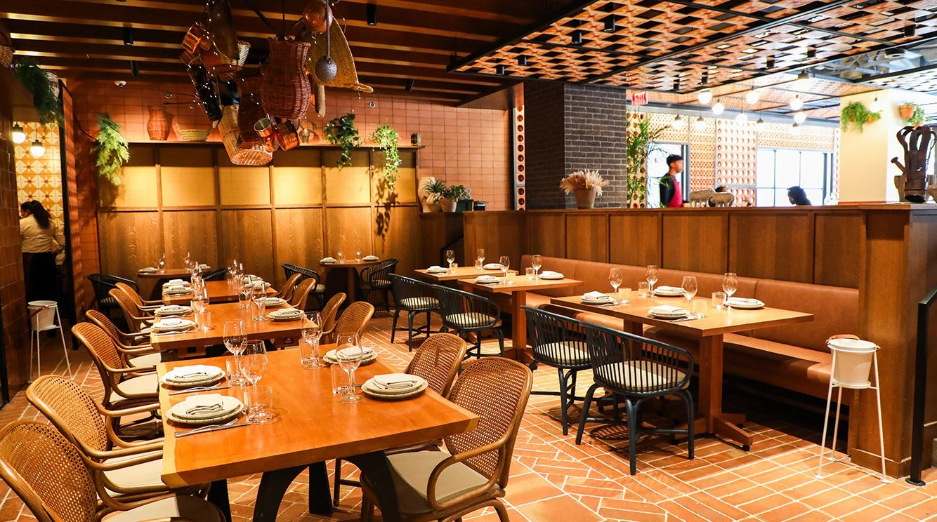 A modern restaurant interior with wooden tables and chairs, setting for dining with plates and glasses, lighted decor, and hanging baskets on the ceiling.