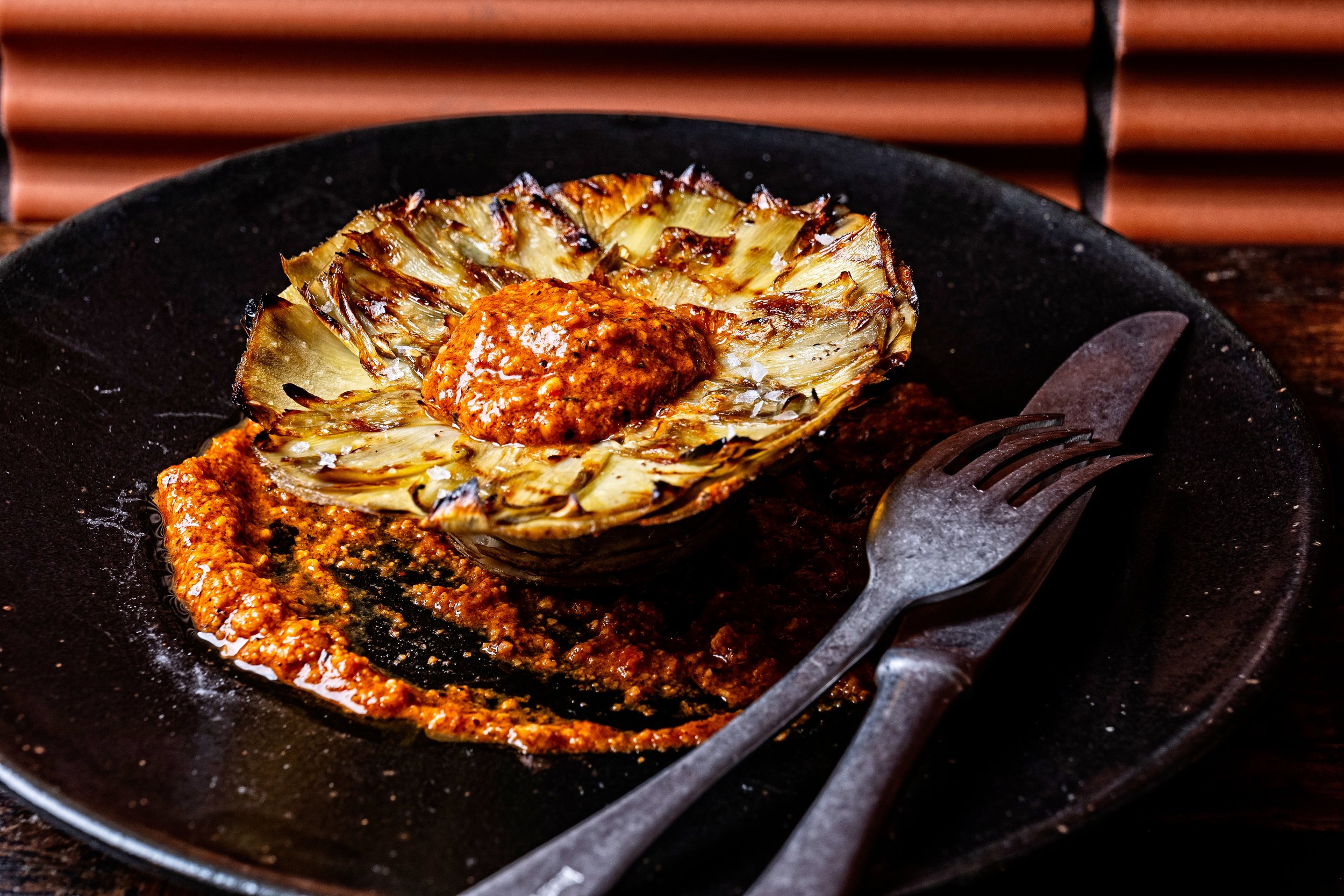 This is an image of a roasting artichoke with a dollop of red sauce on top, placed on a black skillet with a fork and knife nearby.