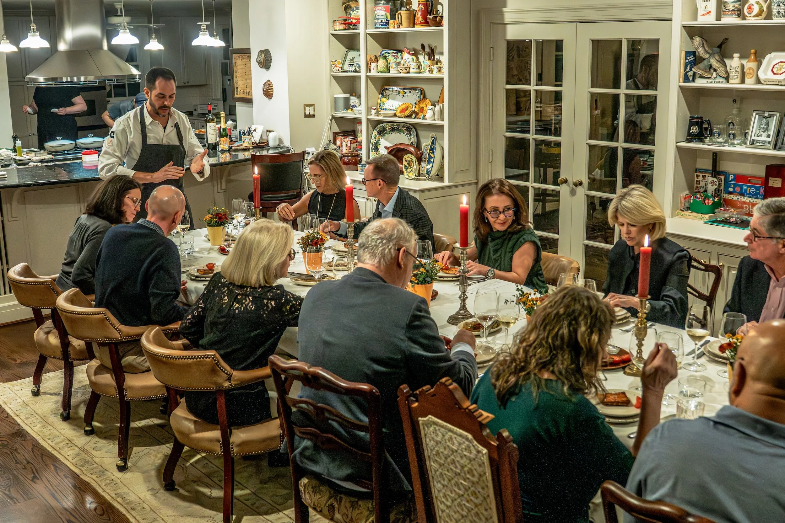 A group of people are seated around a dining table with lit candles, eating dinner in a well-lit, cozy kitchen or dining room decorated with various dishes and collectibles on shelves.