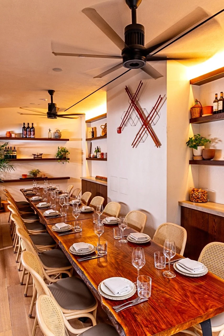 A long wooden dining table set for a meal with plates, glasses, and napkins in a well-lit restaurant with wall shelves and ceiling fans.