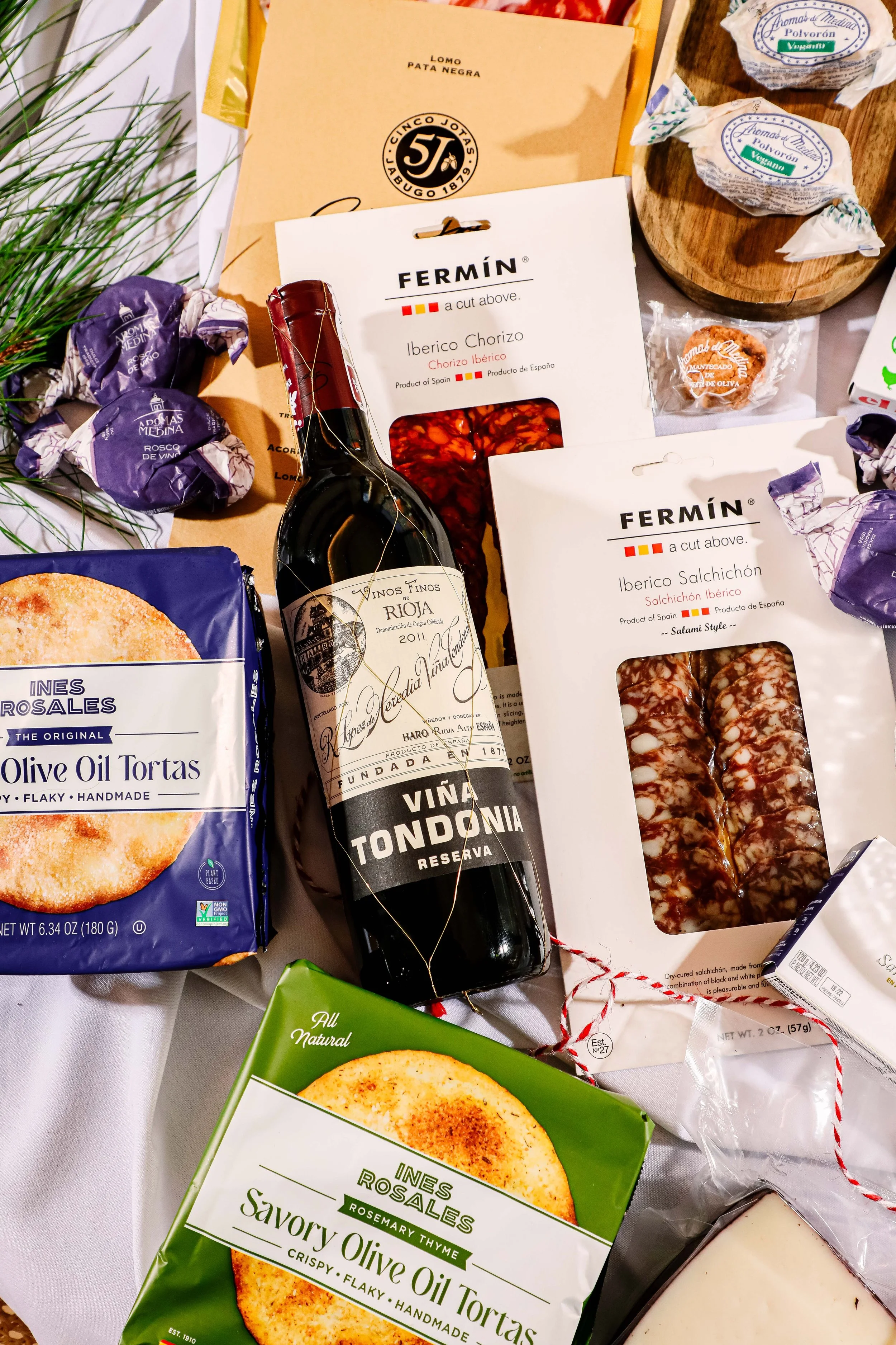 Assorted Spanish food items, including packaged chorizo, salchichón, a bottle of red wine, and packages of olive oil tortas on a white tablecloth.