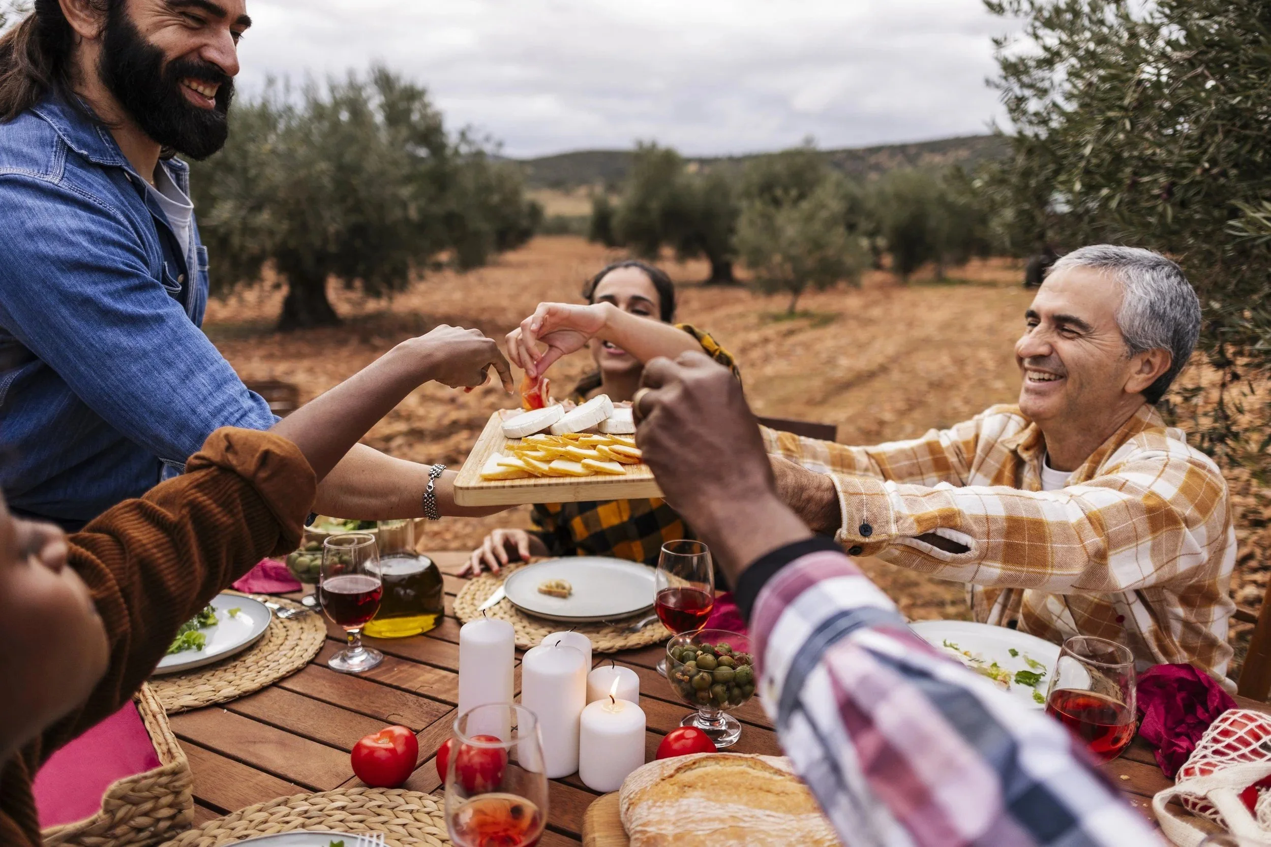 Group of people having outdoor dinner, sharing food, and smiling, with trees and cloudy sky in the background.