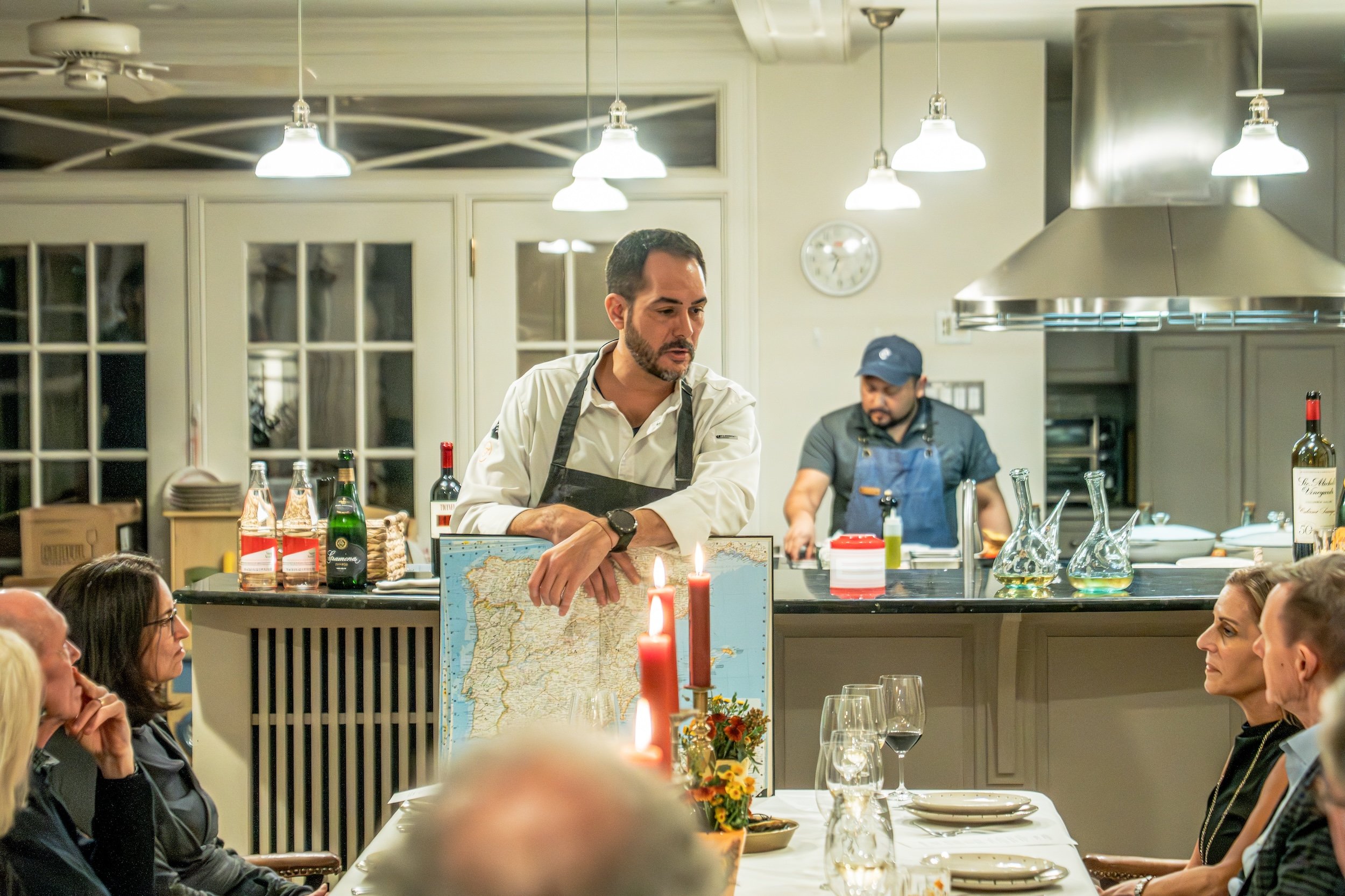 A chef in a white shirt and black apron standing behind a counter with a map of South America, candles, wine bottles, and glasses, preparing food for a group of people seated at a dining table in a restaurant with hanging lights and a fireplace.