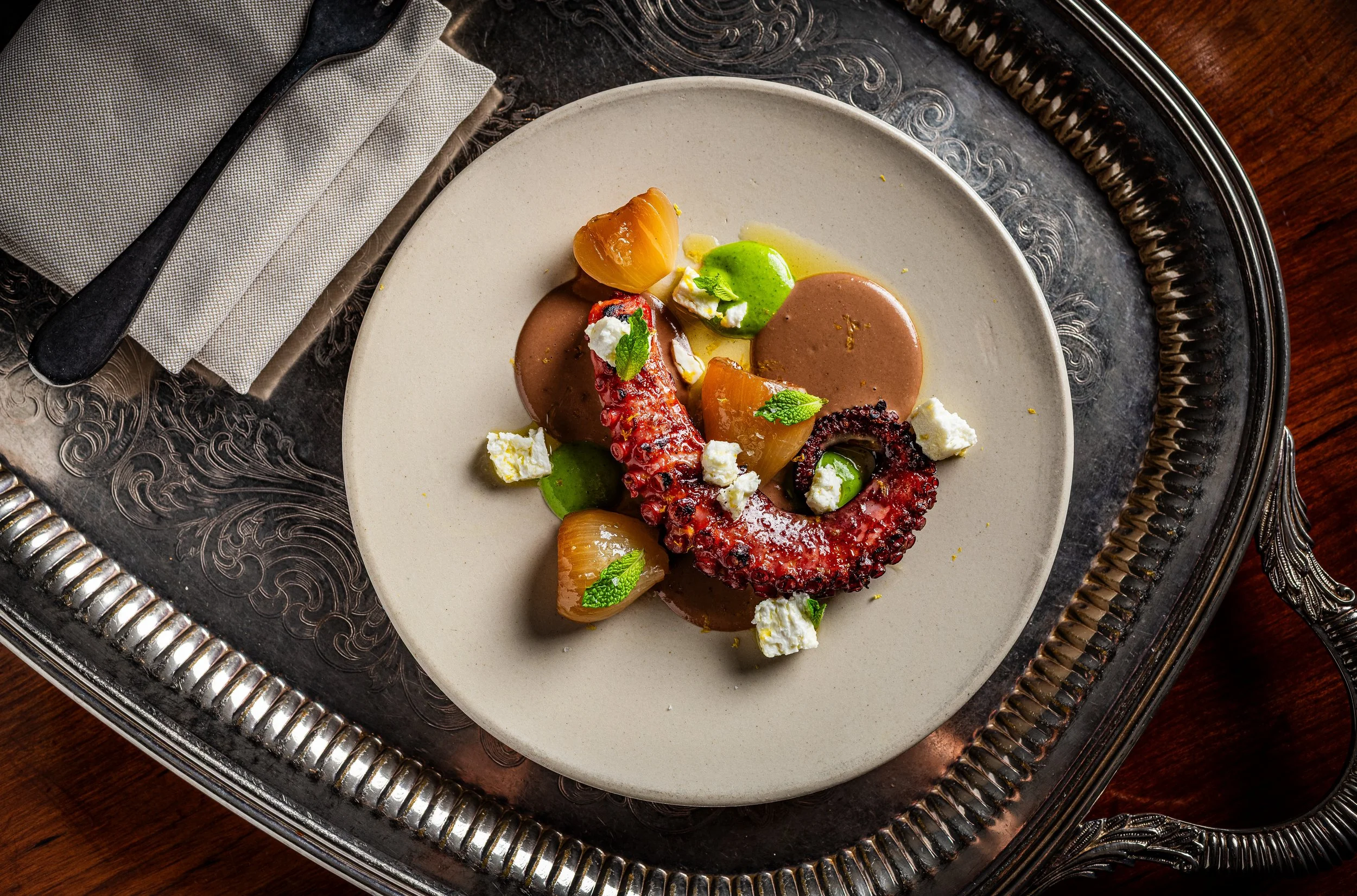 A plate of gourmet seafood dish featuring octopus tentacle, vegetables, microgreens, and sauce, served on an ornate tray.