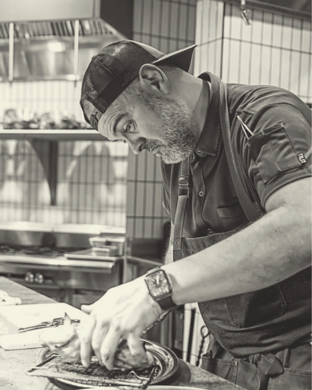 A man working in a kitchen, preparing food, wearing a black shirt, a baseball cap backwards, and a watch.