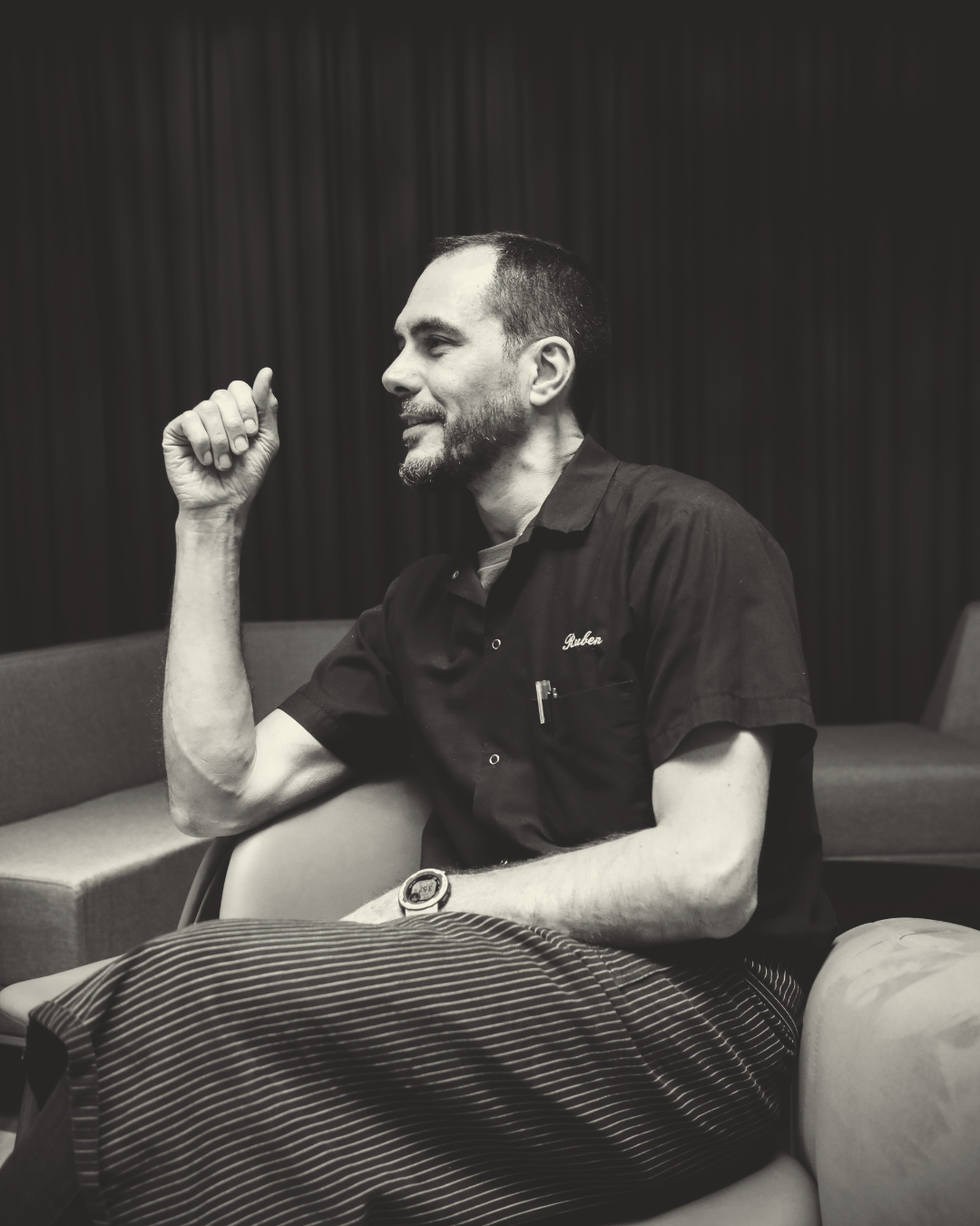 Black and white photo of a man with short hair and a beard, sitting and smiling, wearing a shirt with the name "Ruben" embroidered on it and striped pants, in a room with vertical paneling on the wall.