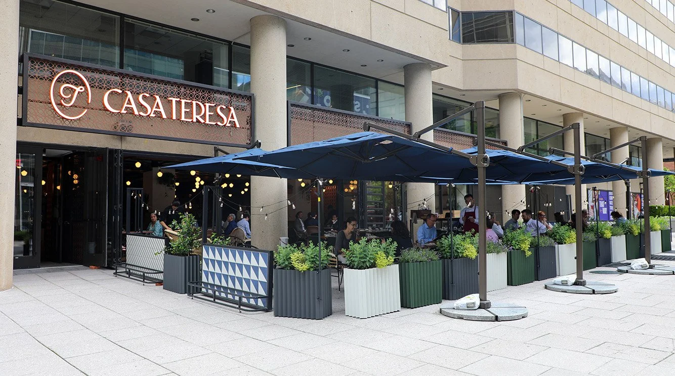 Outdoor seating area of Casatersa restaurant with blue umbrellas, potted plants, and customers dining, in front of a modern building.