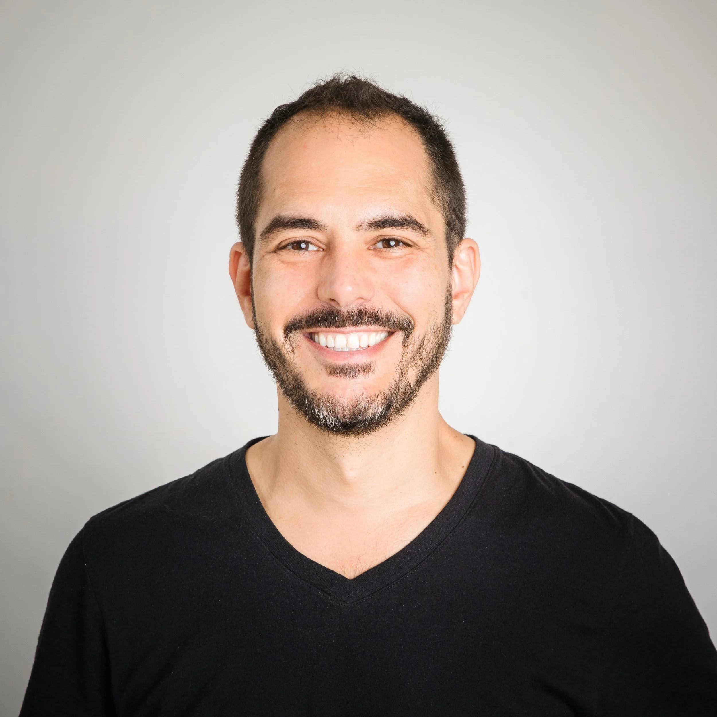 Close-up of a smiling man wearing a black V-neck shirt, with a short beard and dark hair, against a plain light gray background.