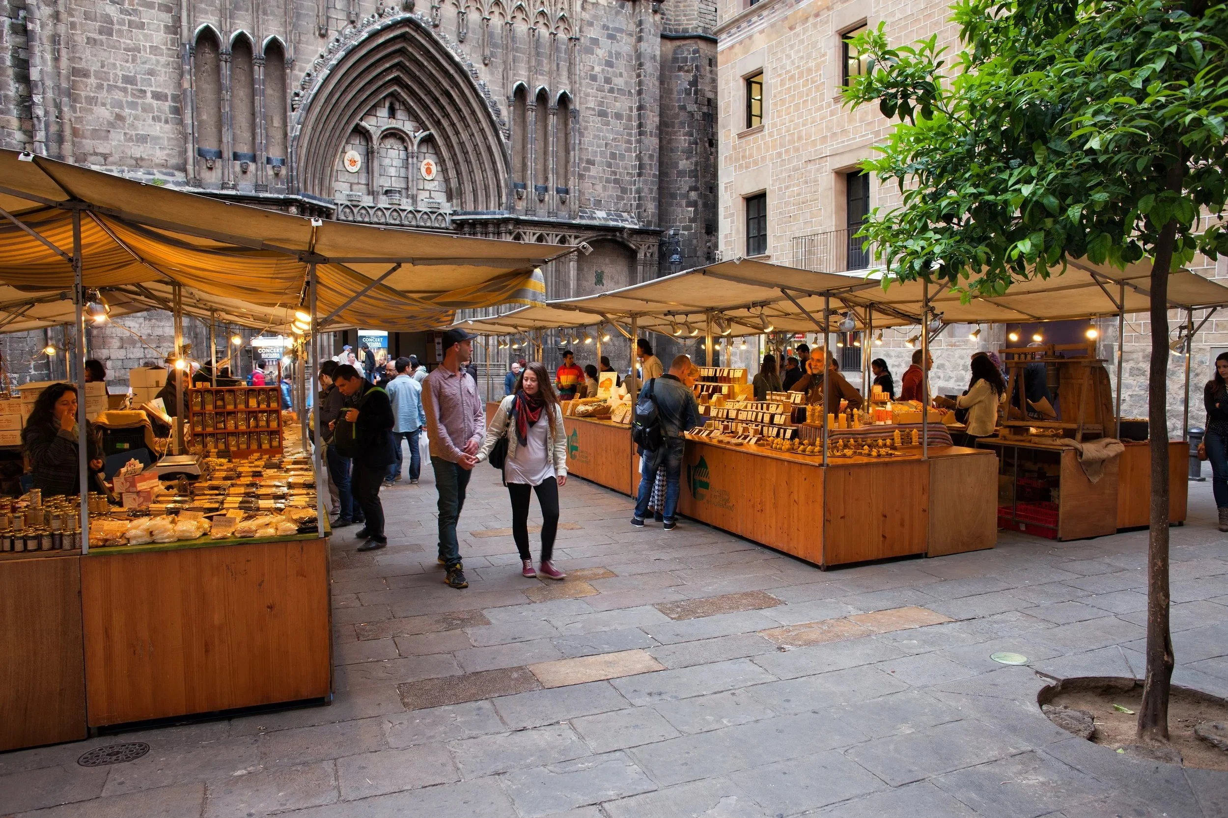 Outdoor market stalls with yellow awnings selling various items, including jewelry and crafts. People browse and walk in front of historic stone buildings with Gothic architecture and a small tree on the right. The area has cobblestone pavement.