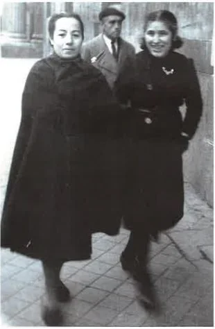 Two women walking together on a city sidewalk with a man in the background.