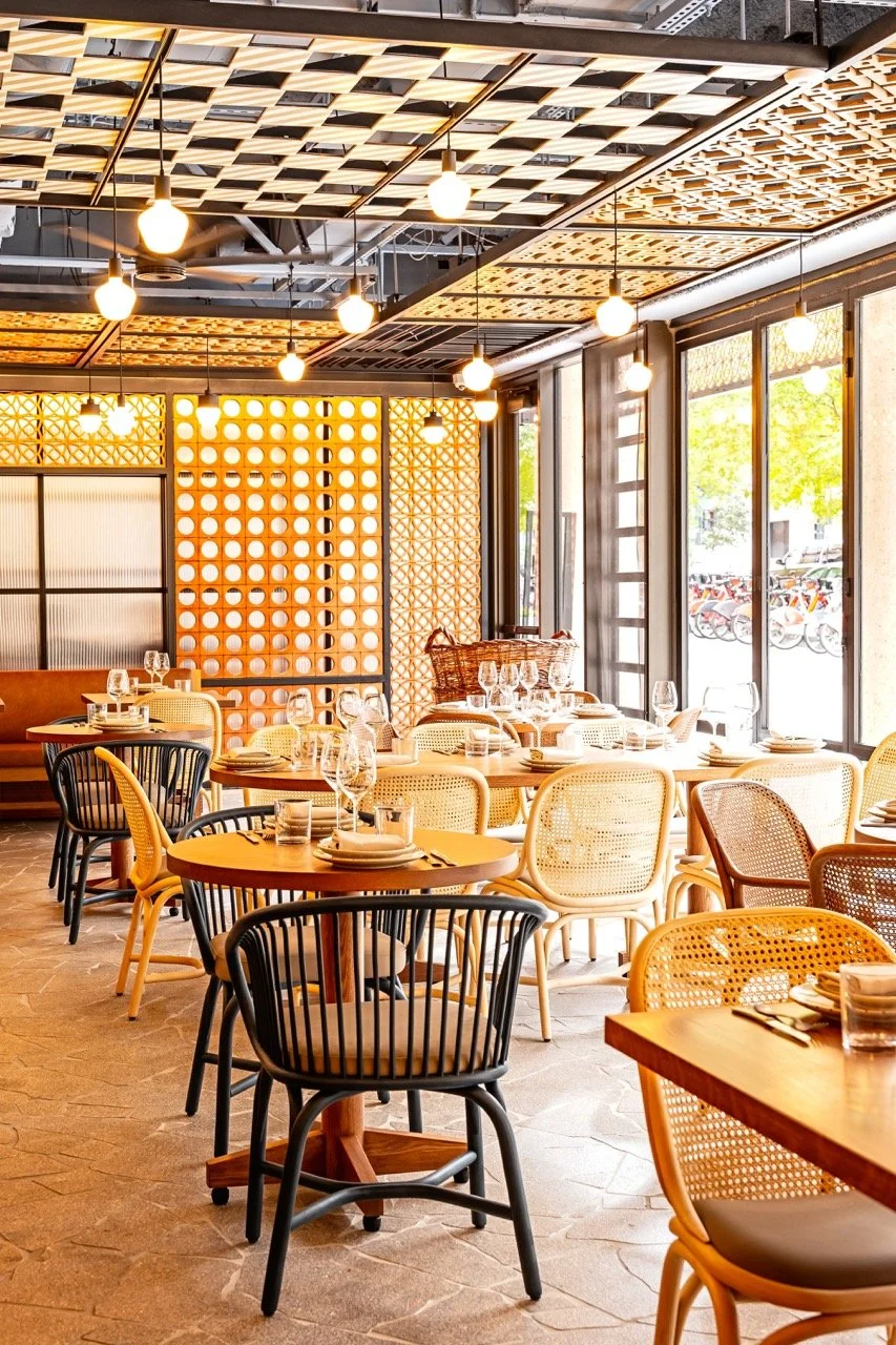 Interior of a modern restaurant with wooden tables and chairs, decorative ceiling panels, hanging light bulbs, large windows with outdoor view of bicycles and trees, and warm ambient lighting.