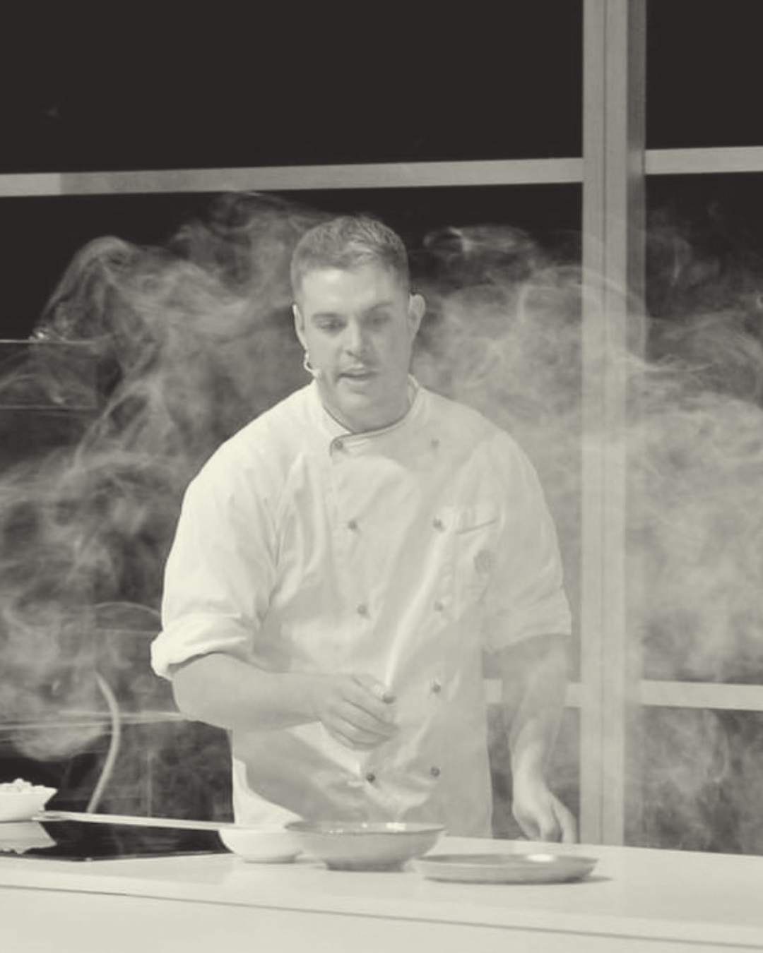 Chef in a white uniform cooking in a kitchen with steam and smoke around him.