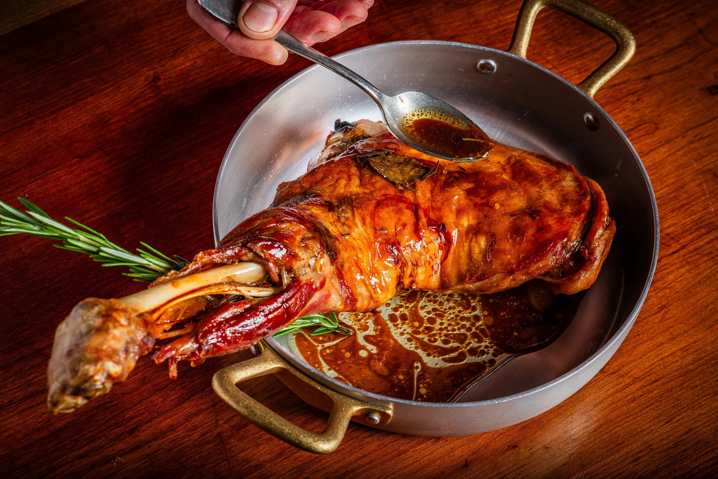 A cooked roast leg of lamb with rosemary, resting in a pan with juices and herbs, on a wooden surface.