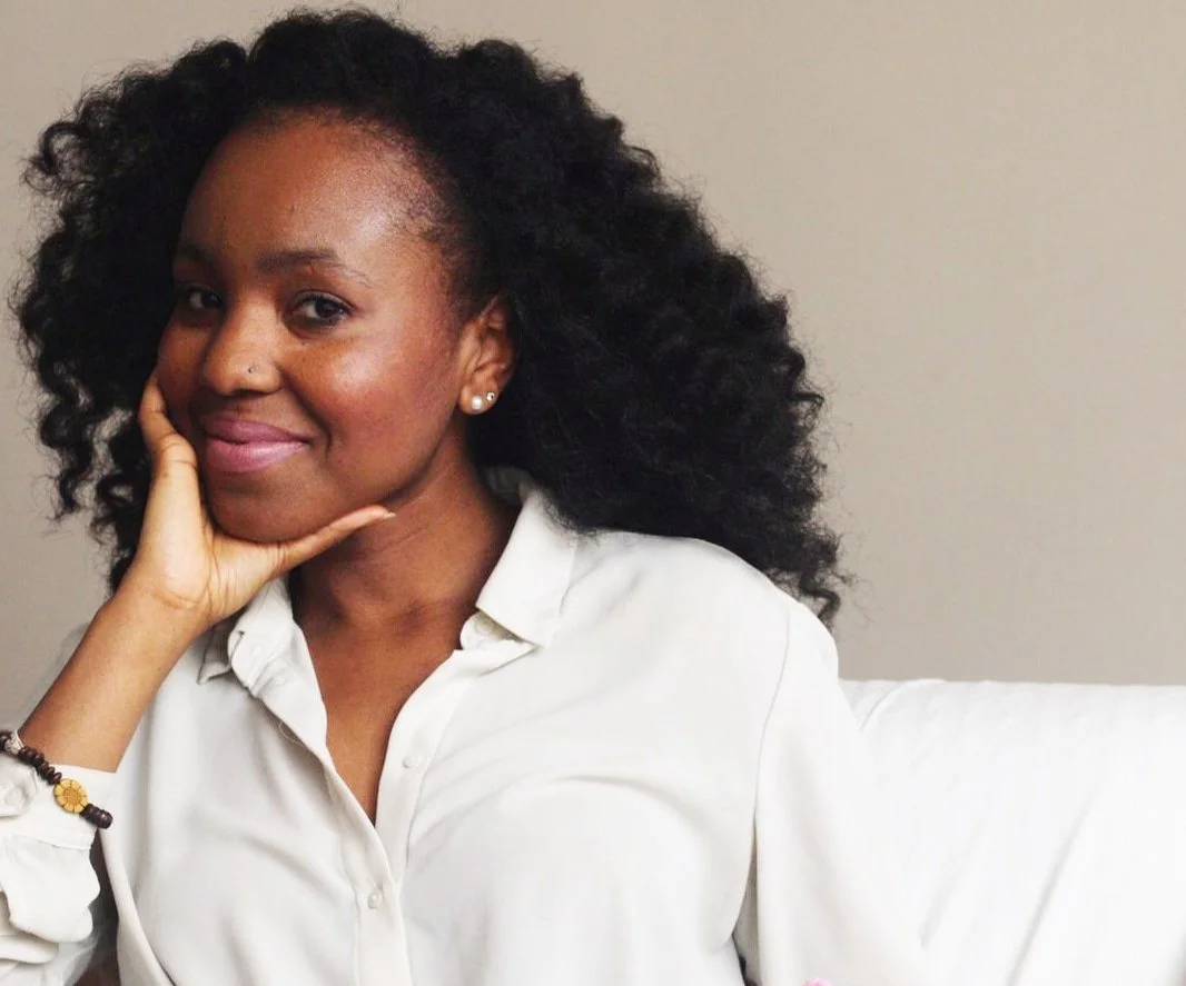A woman with curly black hair, wearing a white blouse, sitting on a white couch, resting her head on her hand, smiling gently at the camera.