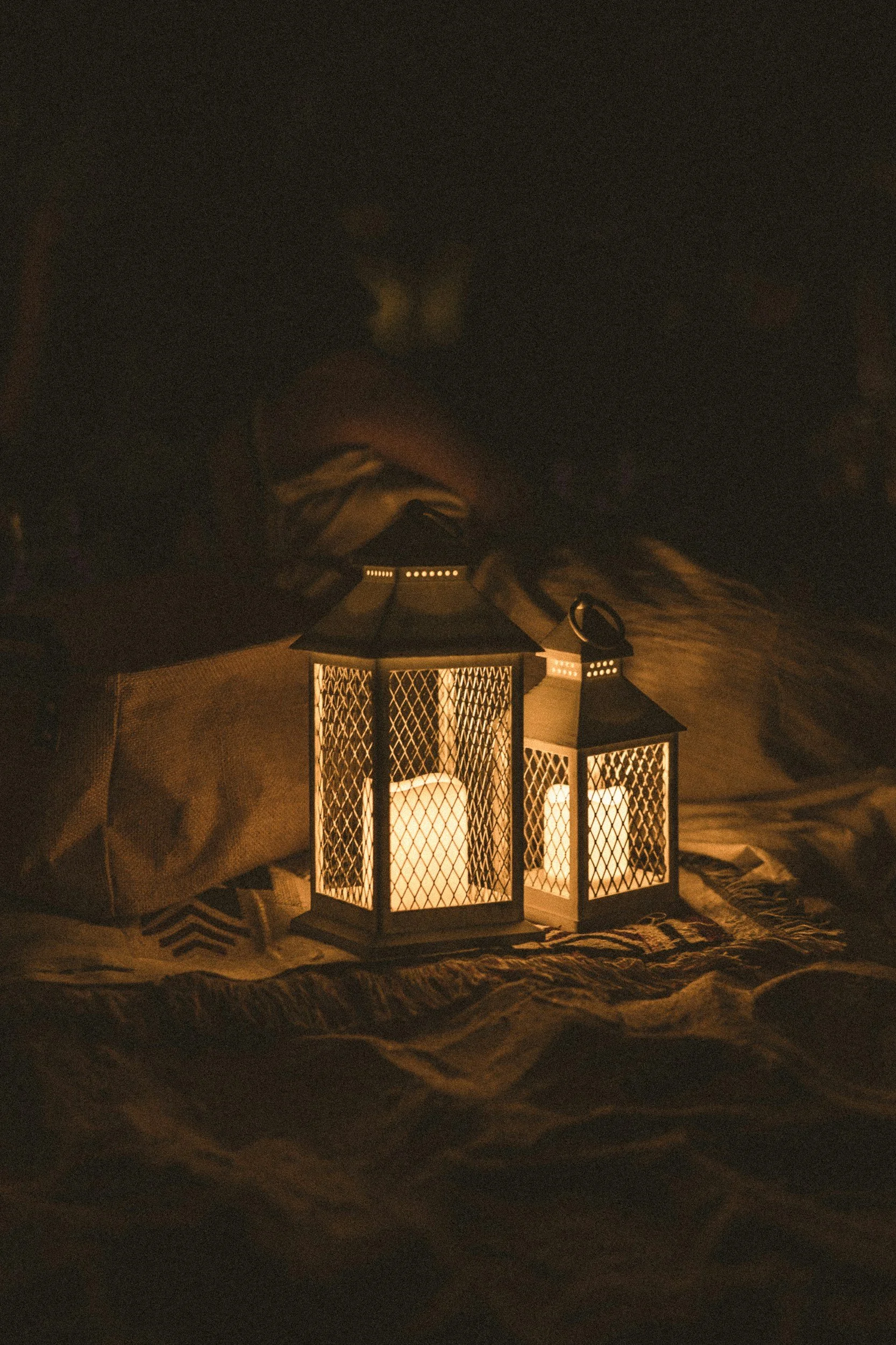 Two lanterns with lit candles inside sit on a fabric surface in a dark setting.