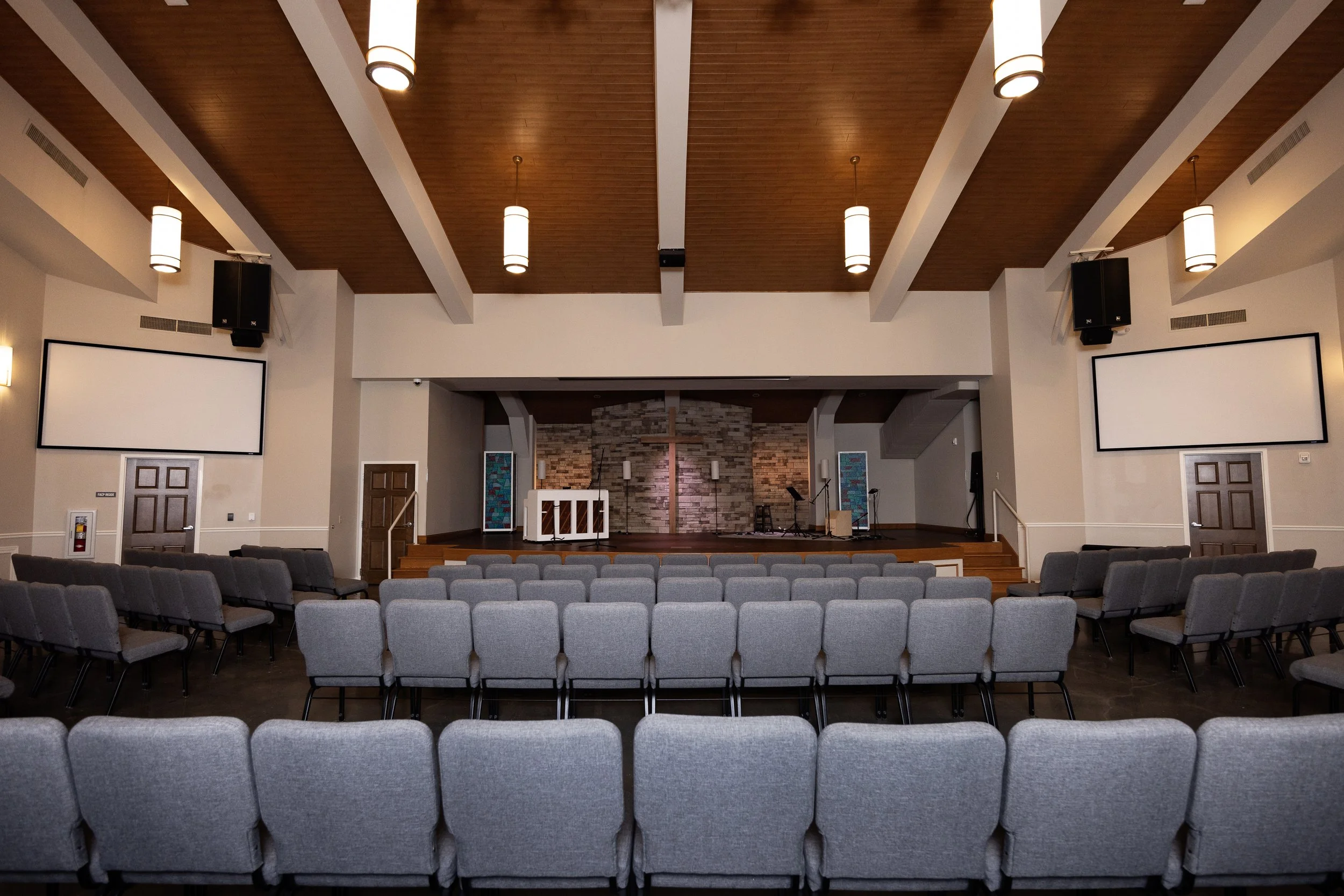 Empty church sanctuary with rows of gray chairs facing a stage with a cross, brick wall backdrop, and musical instruments.
