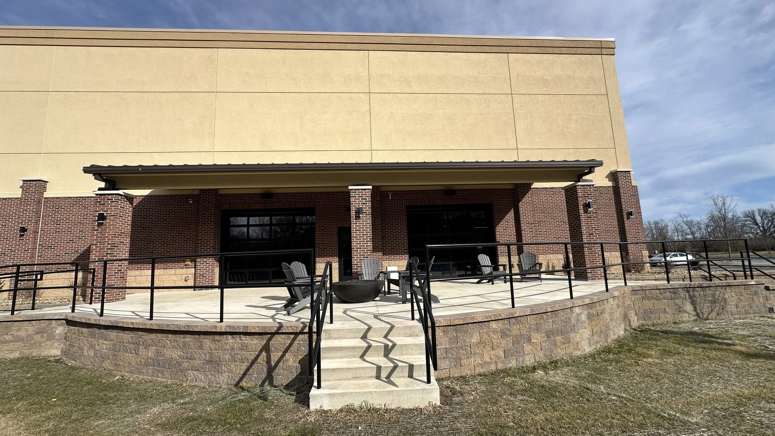 Exterior of a modern building with a brick facade, a concrete terrace with black metal railings, and several black Adirondack chairs. The building has large glass windows and doors, a sloped roof, and a beige upper wall. Steps lead up to the terrace,