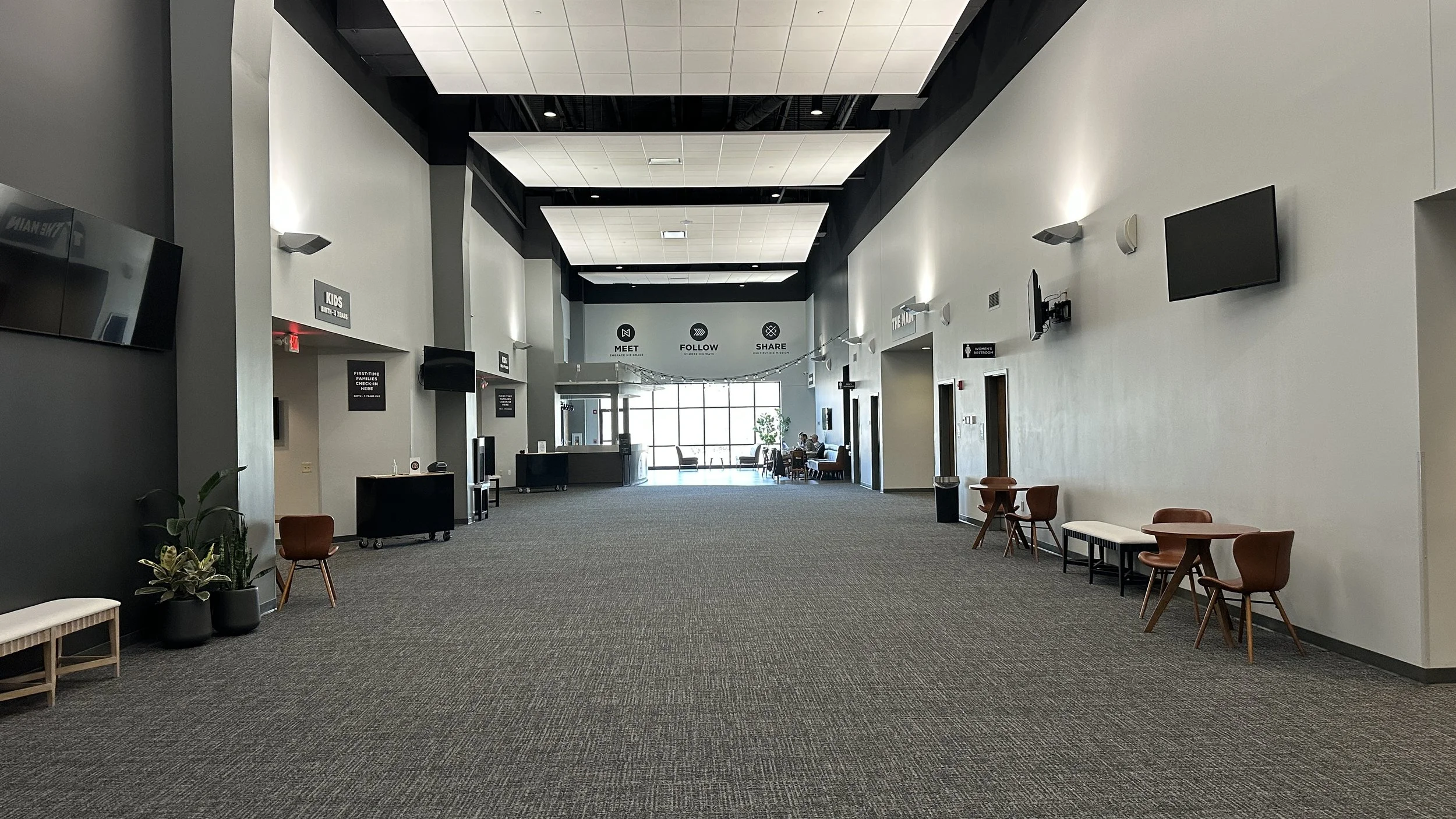 A spacious, modern indoor lobby with high ceilings and neutral-colored walls. There are chairs and small tables along the walls, with a few people sitting near the large windows at the back. Wall-mounted TVs and signs are visible, and the floor is ca