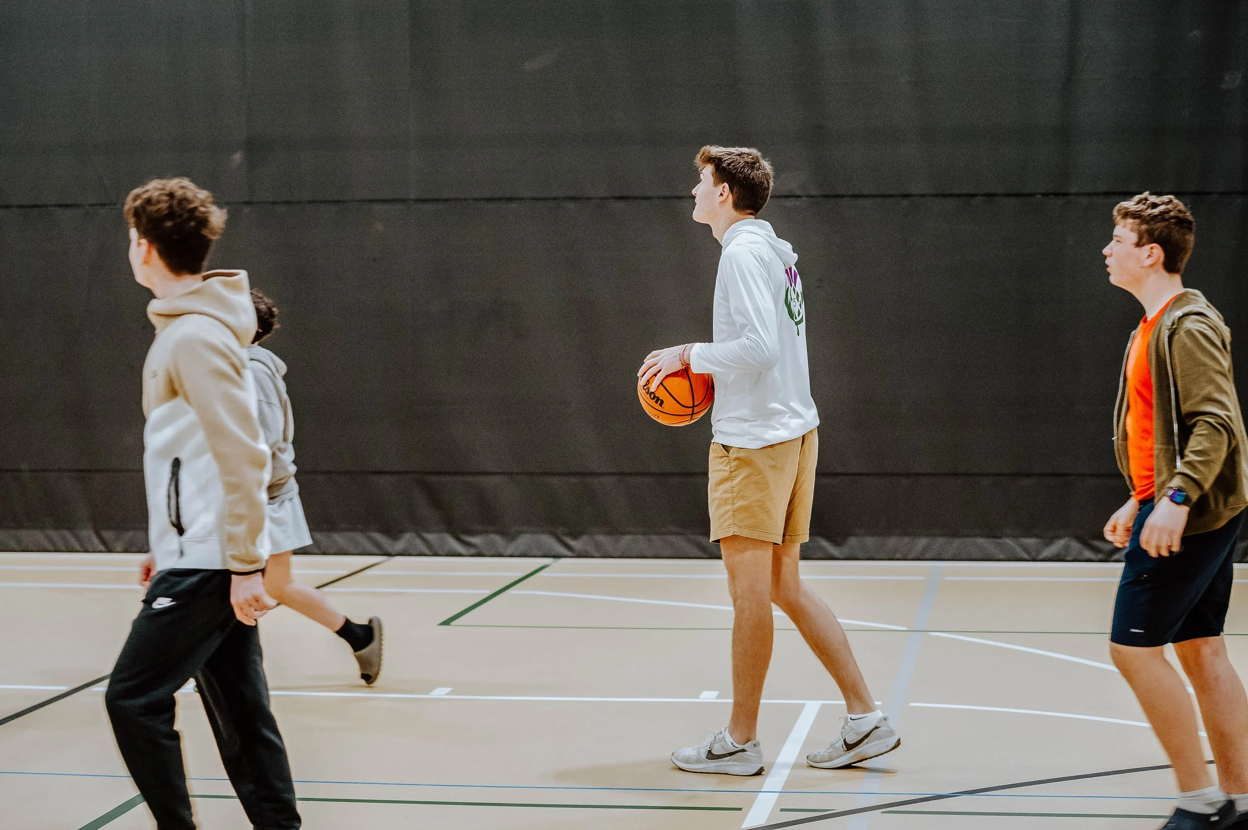 Group of young males in casual clothes playing basketball in an indoor court.