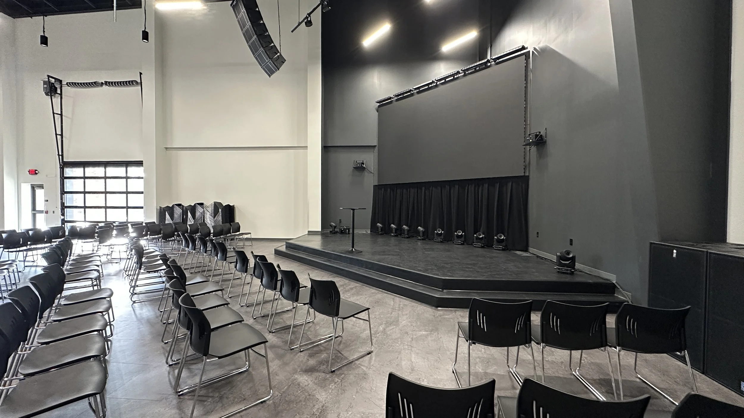 Empty conference room with rows of black chairs, a stage with a microphone stand, and a large screen, with stage lights and equipment, in a modern building with high ceilings and large windows.