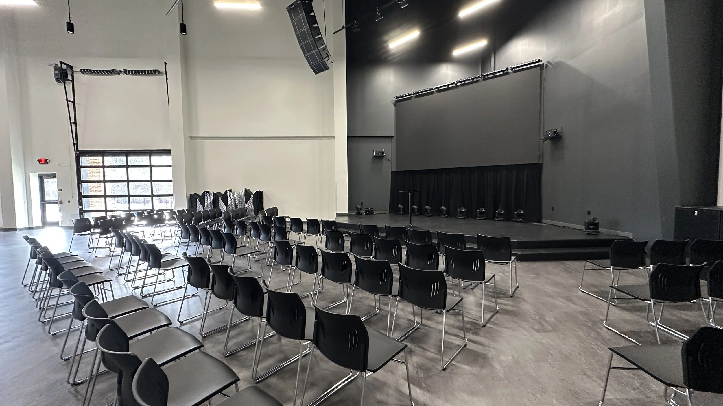 Empty event space or auditorium with rows of black chairs facing a stage with a black curtain, equipped with stage lights, speakers, and a microphone stand.