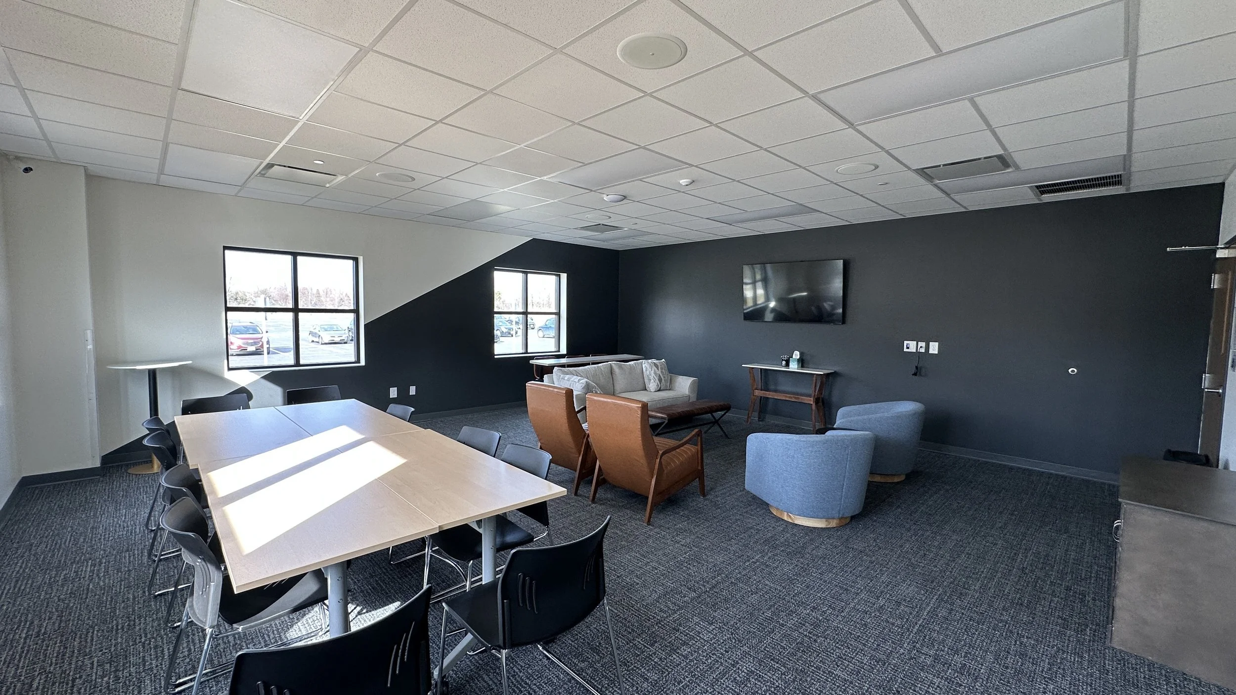 Interior view of a waiting or meeting room with a large rectangular table and multiple chairs, a cozy seating area with a sofa and armchairs, a wall-mounted flat-screen TV, and windows providing natural light.