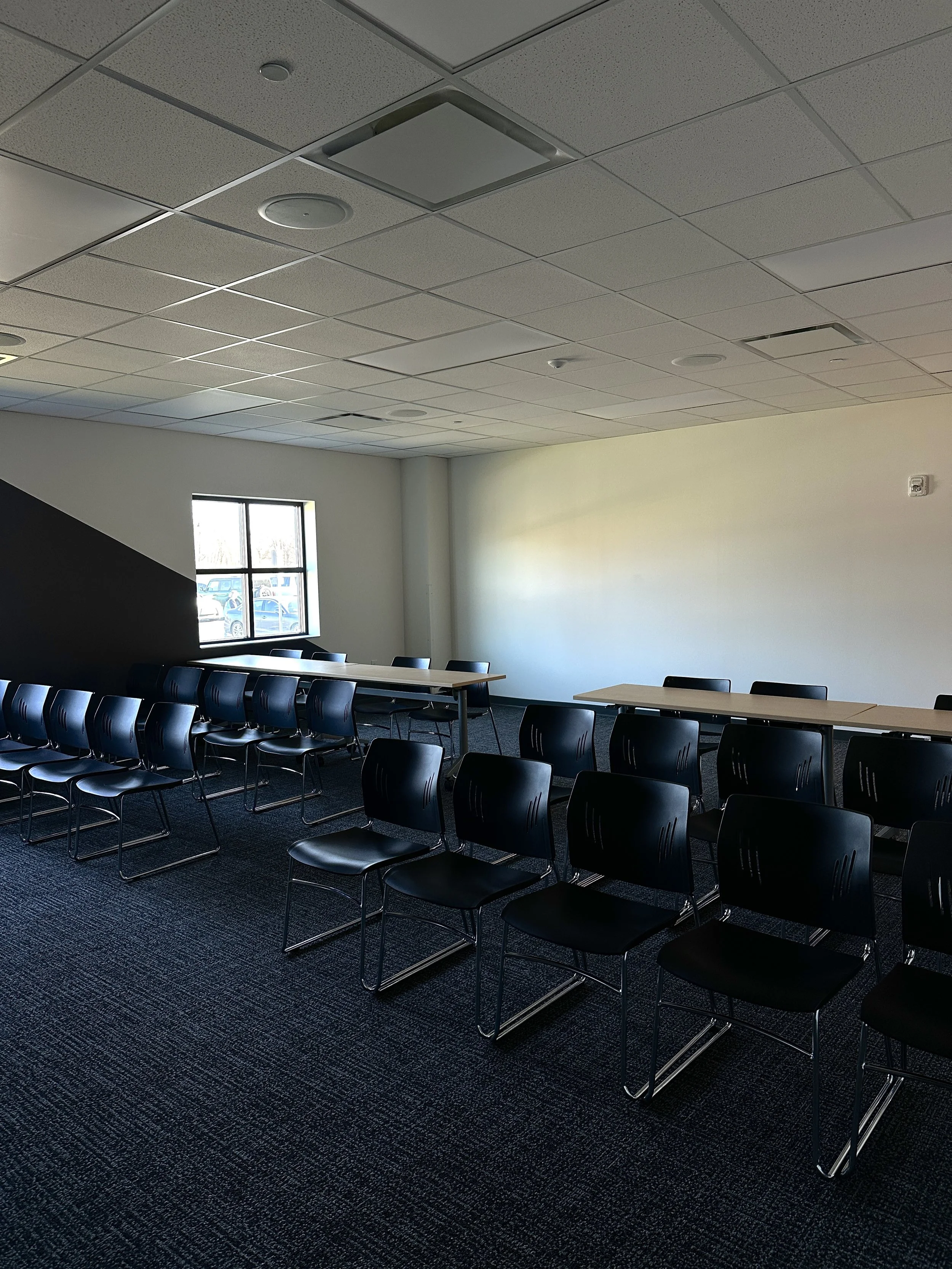 Empty room with black chairs arranged in rows, tables along the wall, and a window letting in natural light.