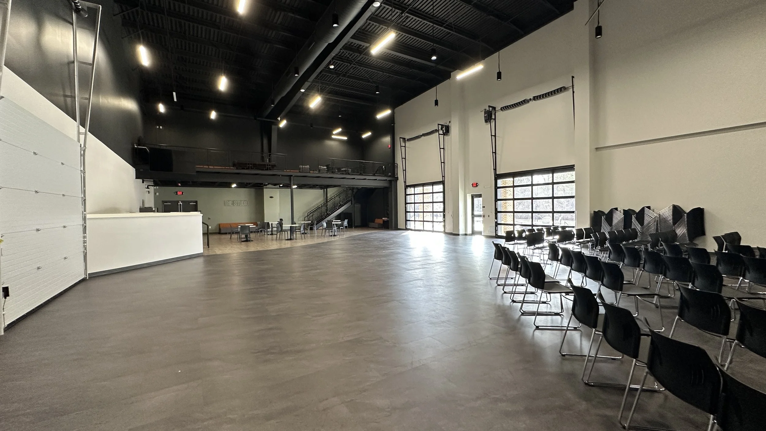 Empty event space with black chairs arranged on the right, multiple large windows on the right wall, and a small tiered seating area with tables and chairs in the back. The front area is open and spacious with a high ceiling.