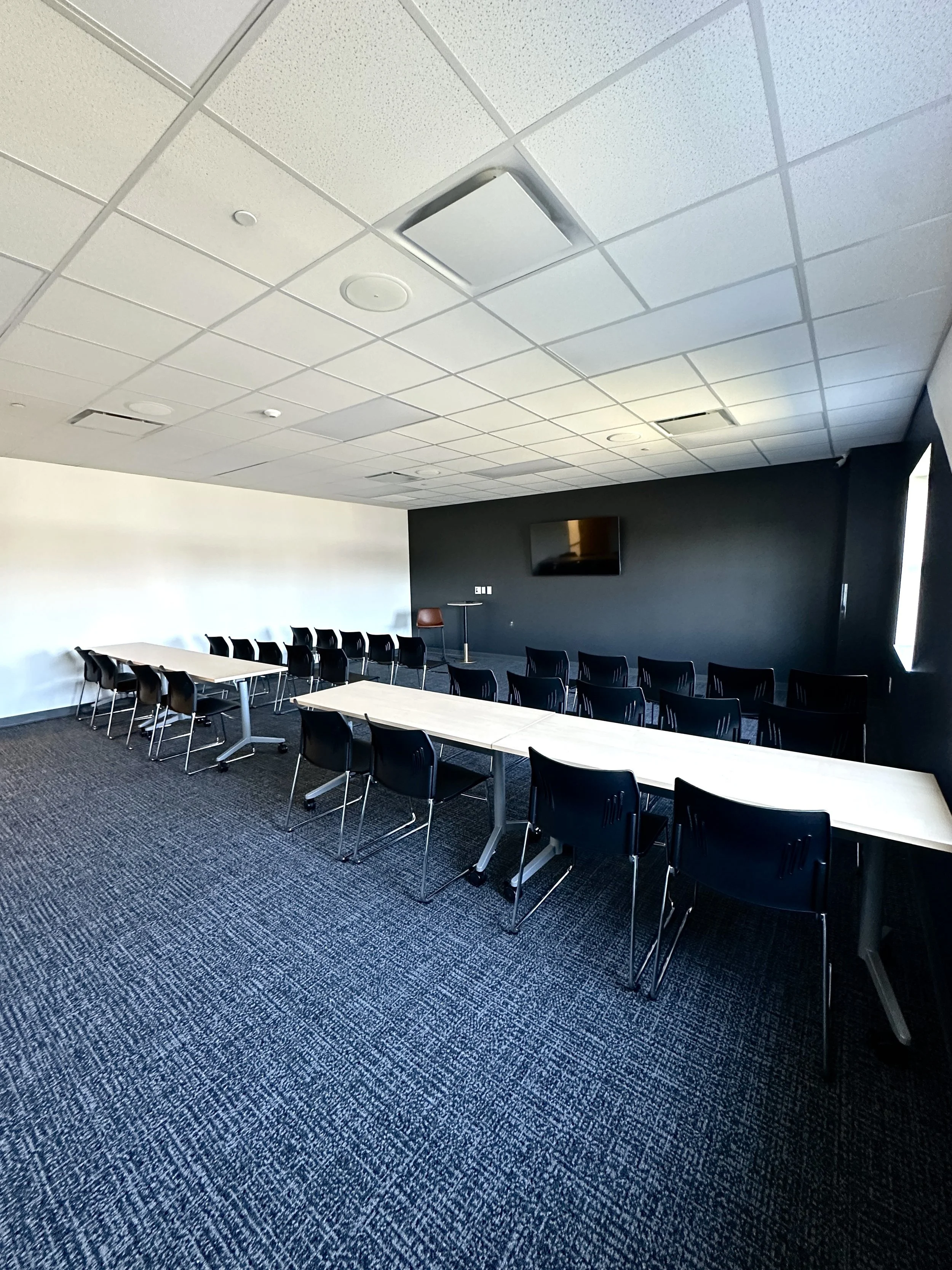 Empty conference room with long white tables, black chairs, a black wall with a mounted TV, and a small standing table with a brown chair. Carpeted floor and a window on the right letting in natural light.