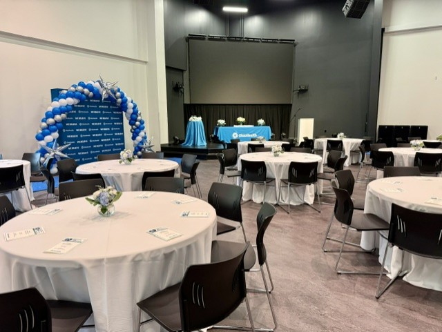 Event space set up with round tables covered with white tablecloths, decorated for a celebration. Blue balloon arch and a table with flowers, with a blue and white theme. Stage in the background with a blue tablecloth and floral arrangements.