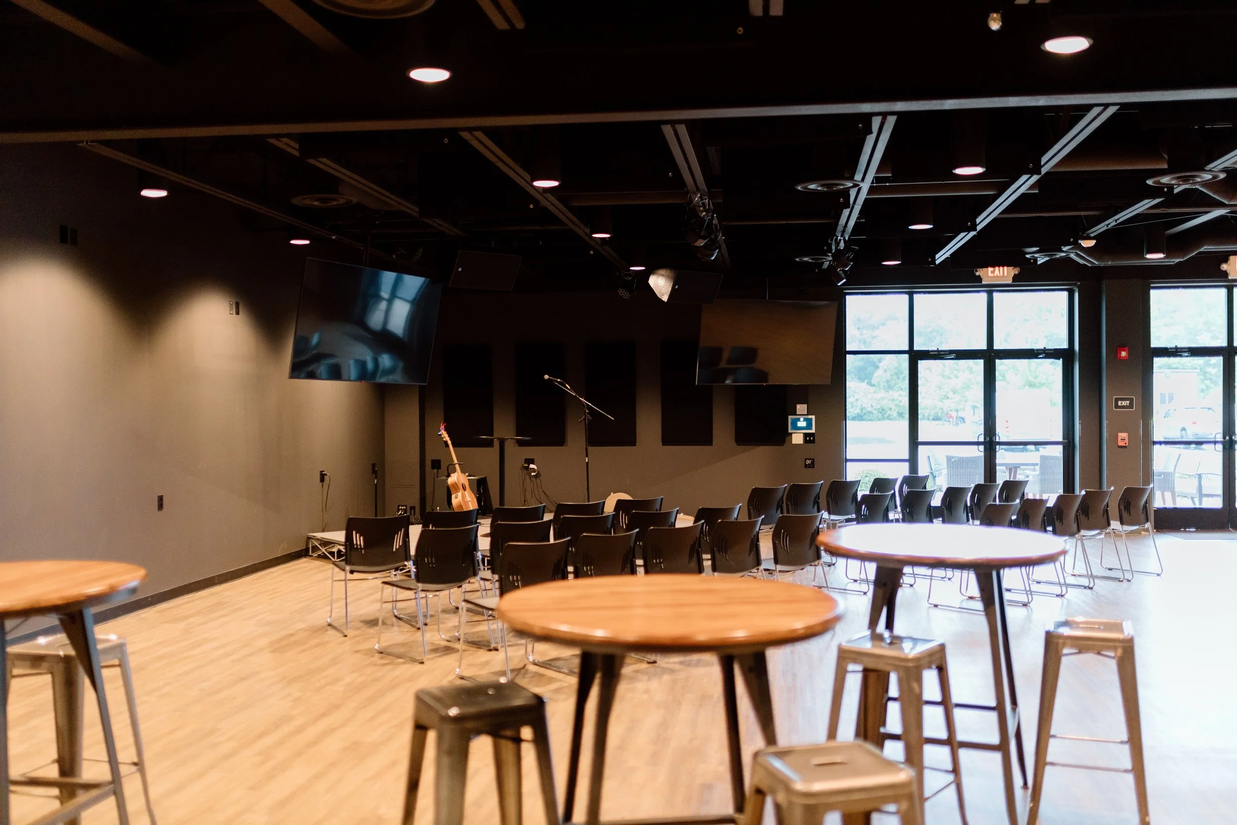 Empty stage with guitars, microphones, and monitors in a modern, dimly lit performance venue with wooden floors, large TVs, and glass doors letting in natural light.