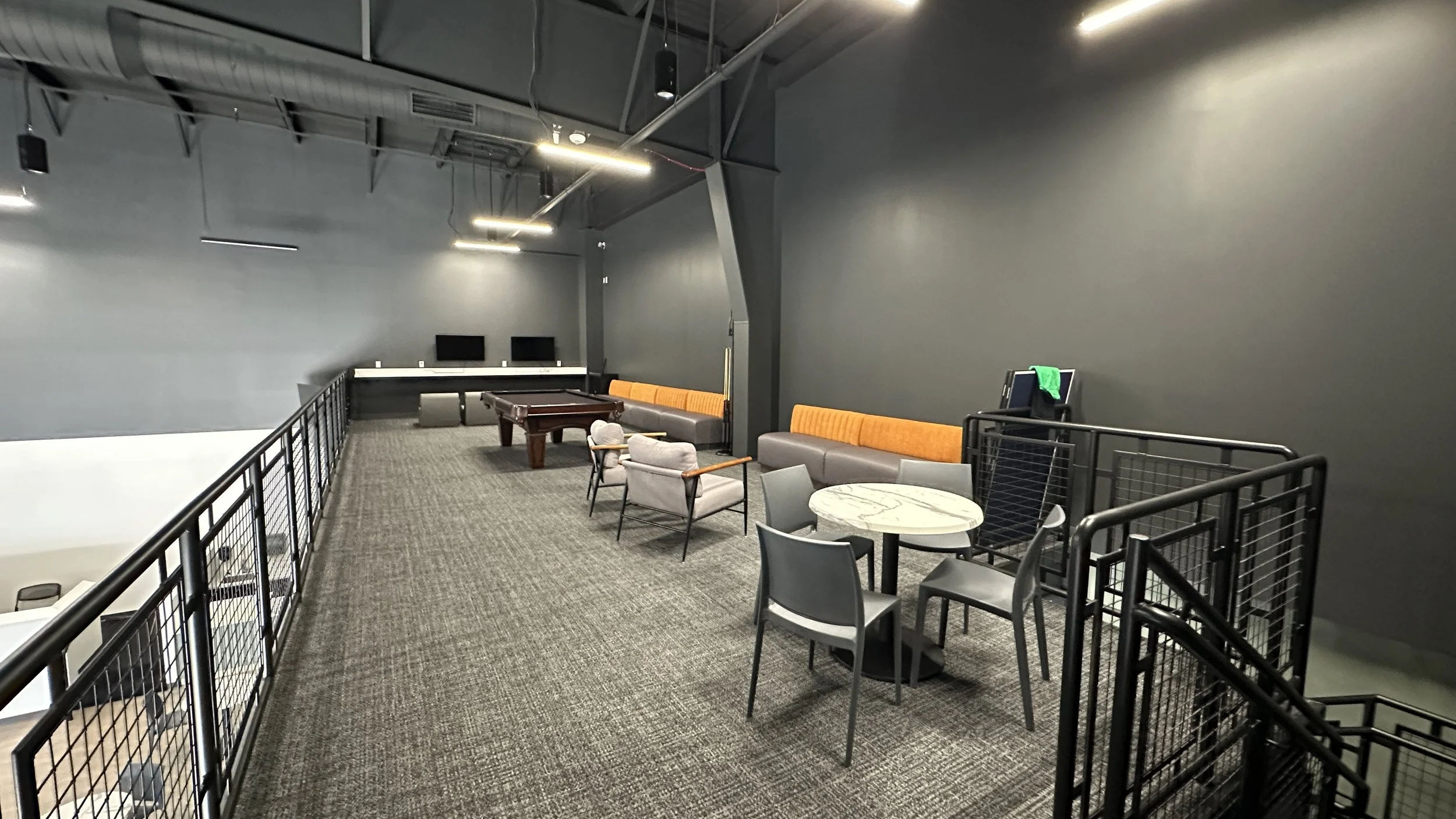 Interior of a modern lounge area with gray walls, a gray carpet, and black ceiling beams. There are various seating options including gray, orange, and patterned chairs and couches, a small marble-top table, a pool table, two computer monitors on a c