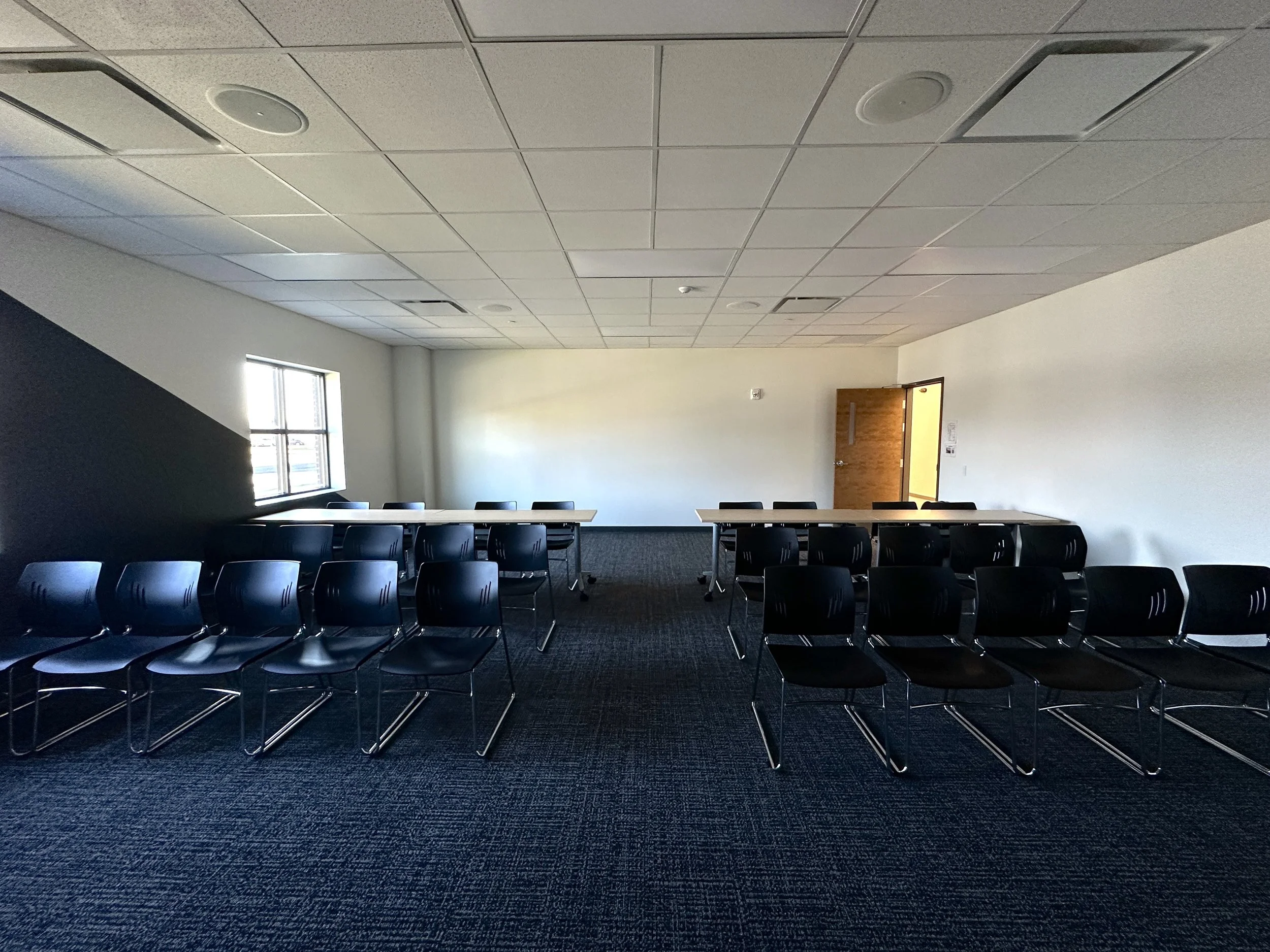 Empty conference room with black chairs arranged in rows, tables, a window on the left wall, and a door at the back.