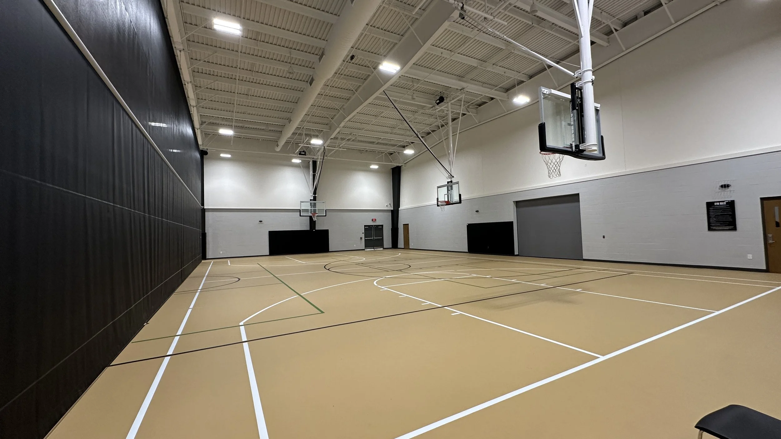 An indoor basketball gymnasium with three basketball hoops, a tan floor with white, black, and green lines, and a high ceiling with lights.