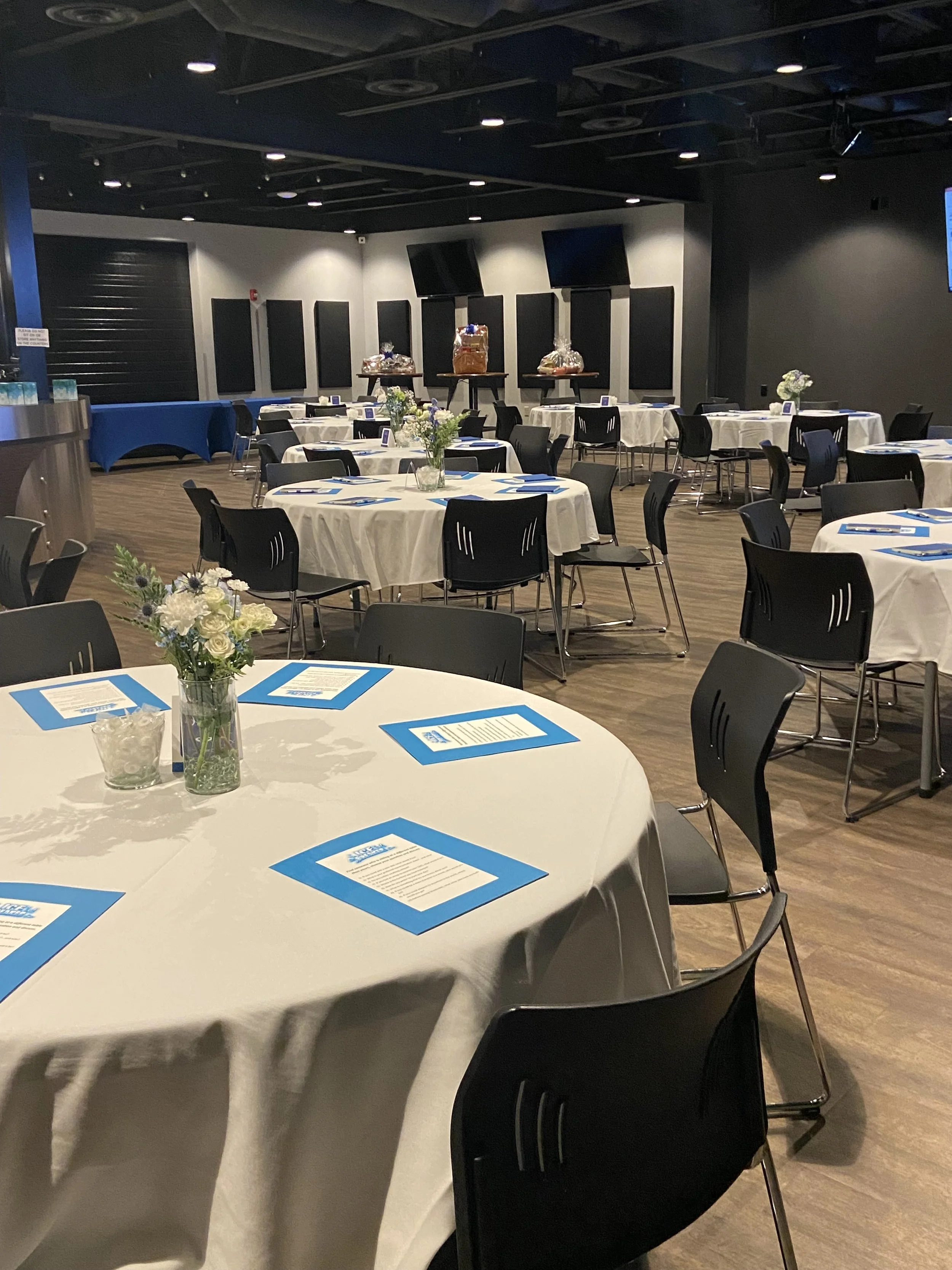 A banquet hall with round tables covered with white tablecloths, decorated with small flower arrangements, and blue and white printed menus. Chairs surround the tables, and a buffet area with gift baskets is visible in the background.