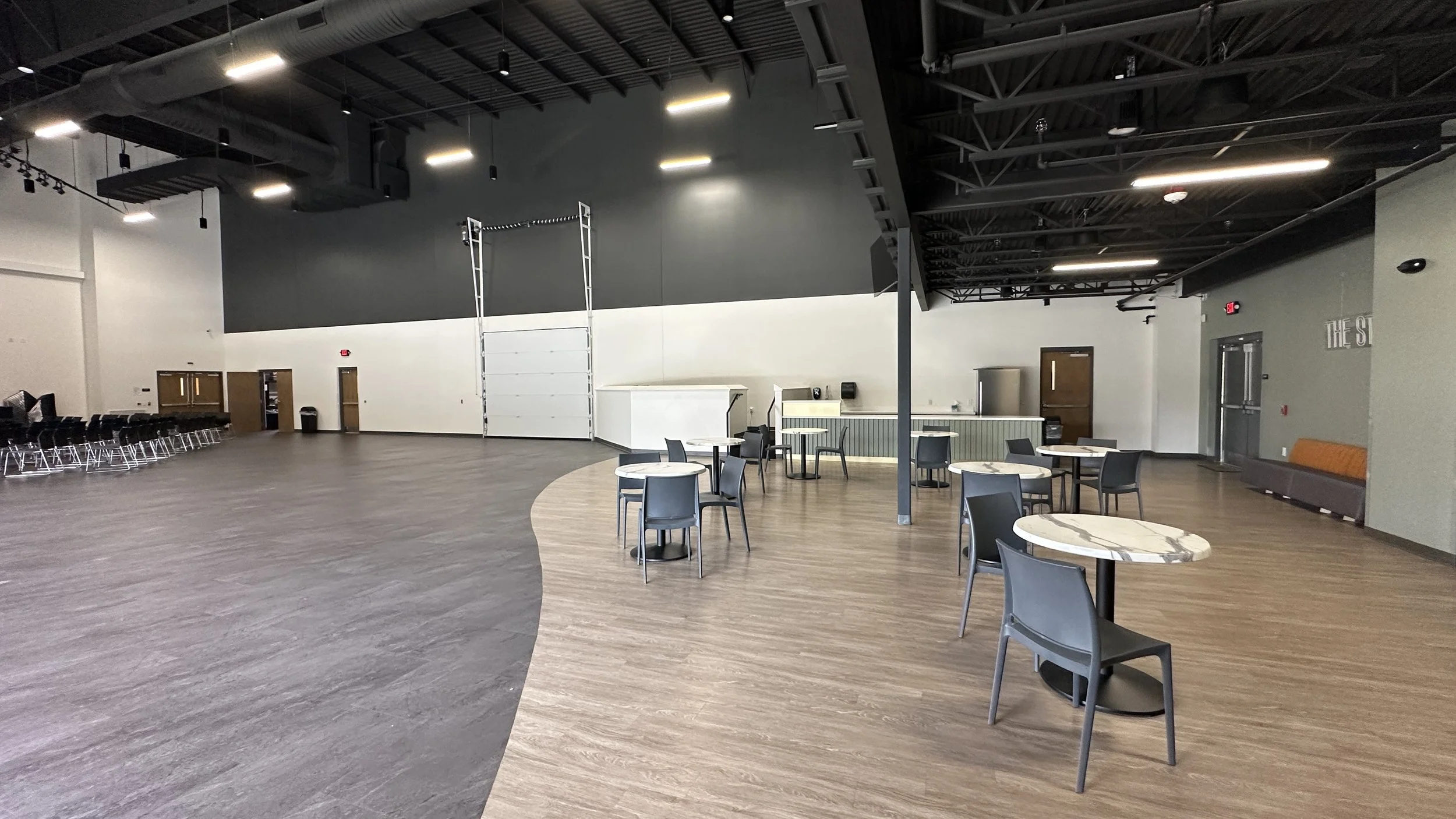 Spacious indoor area with a curved wooden floor section, scattered round tables with chairs, black chairs, a small kitchen counter, and a waiting area with chairs and a bench, in a modern venue with high ceilings and black and white walls.