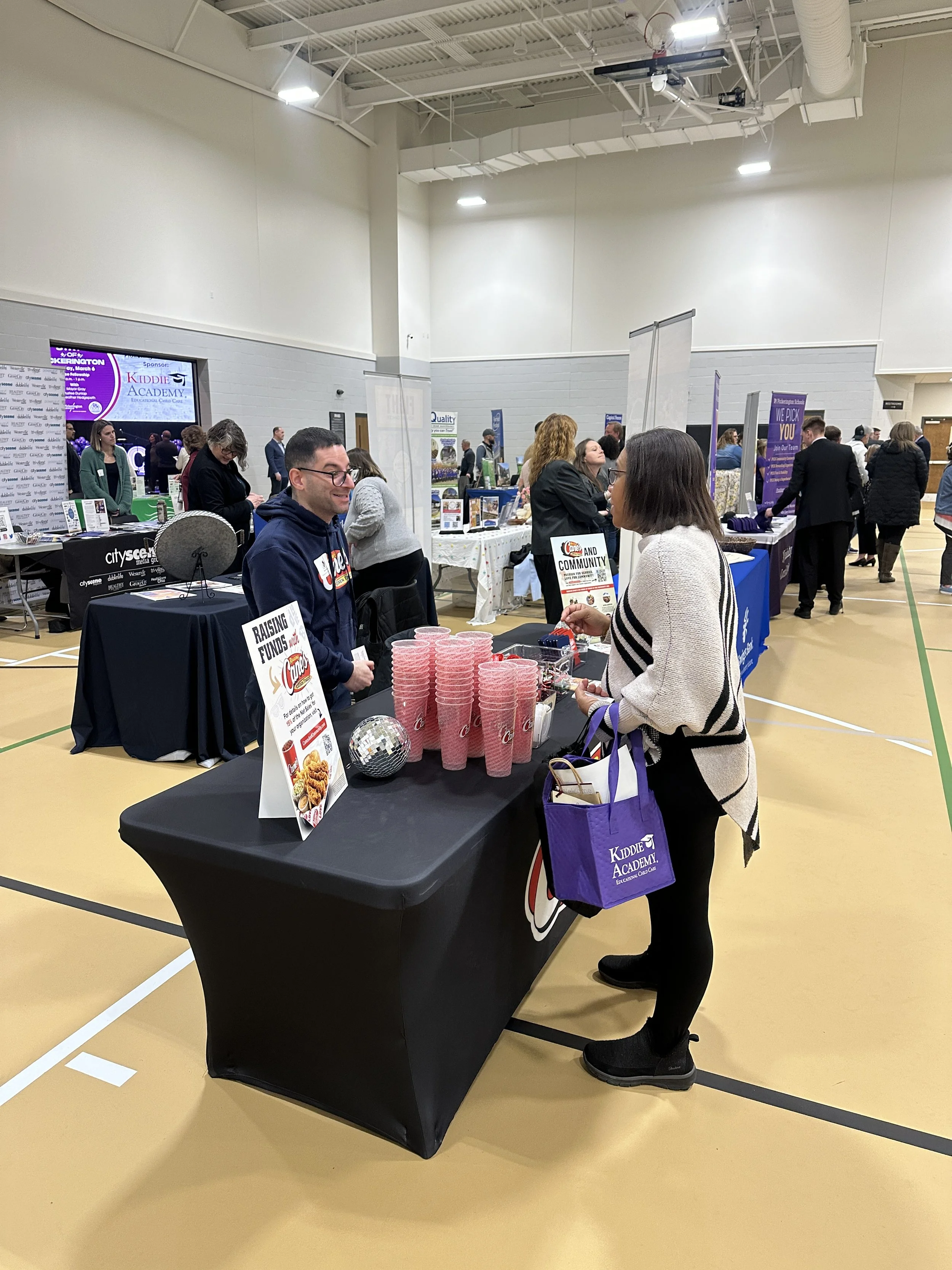 A woman engaging with a man at a booth during an indoor event or fair, with several other booths and attendees in the background. The woman is holding a purple Kidde Academy bag. The booth has multiple pink cups and signage.