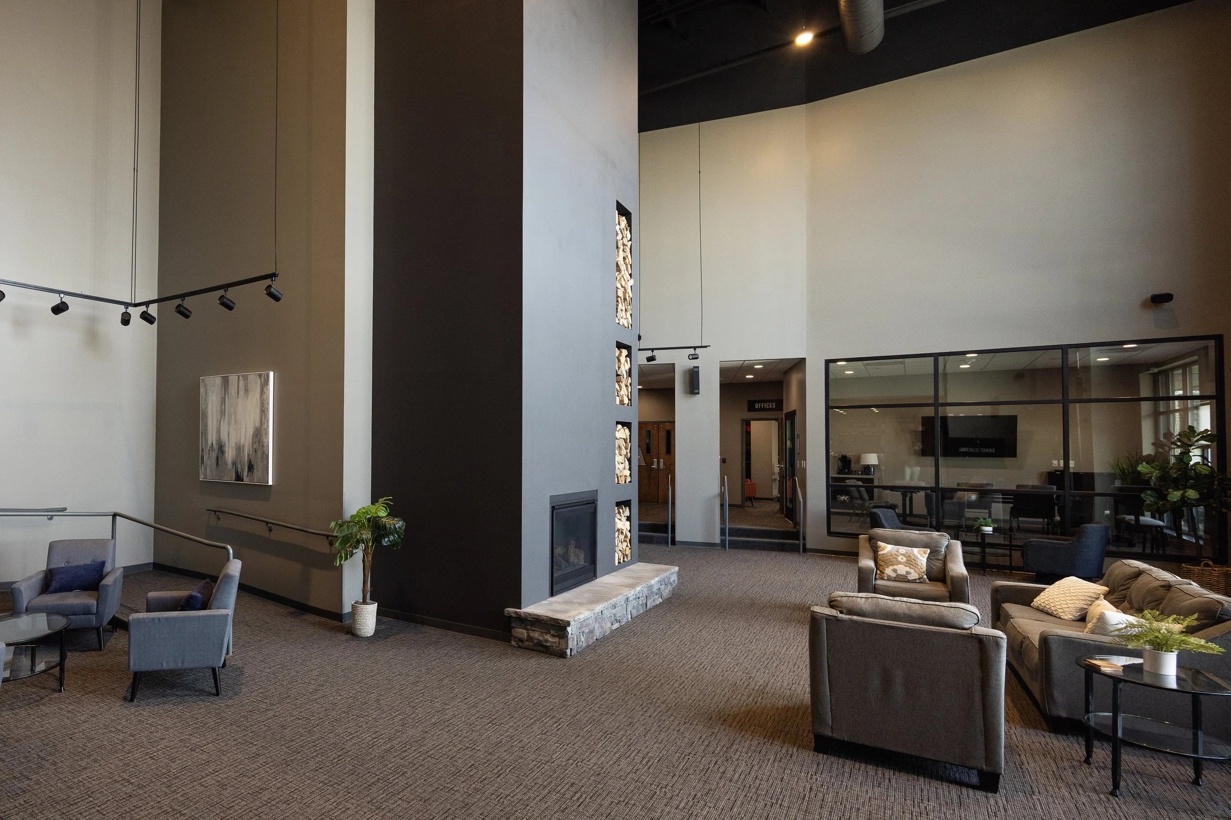 Modern office lobby with seating area, a black fireplace with firewood, and a glass-walled conference room, decorated with houseplants and artwork.