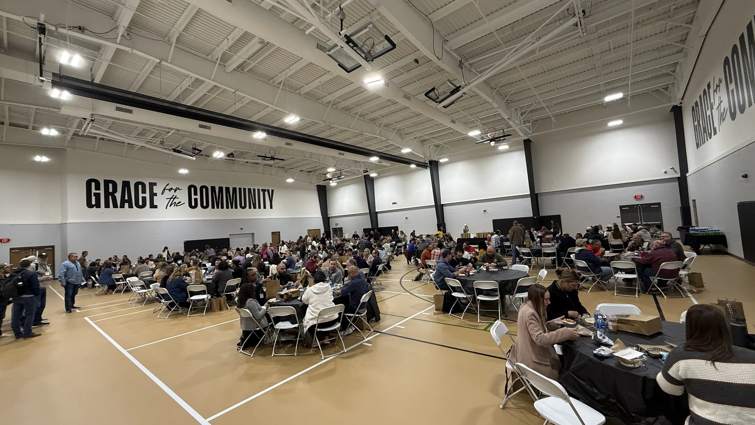 Large indoor community event in a gymnasium with many round tables and chairs, people eating and socializing, with a sign on the wall that says 'Grace for the Community'.