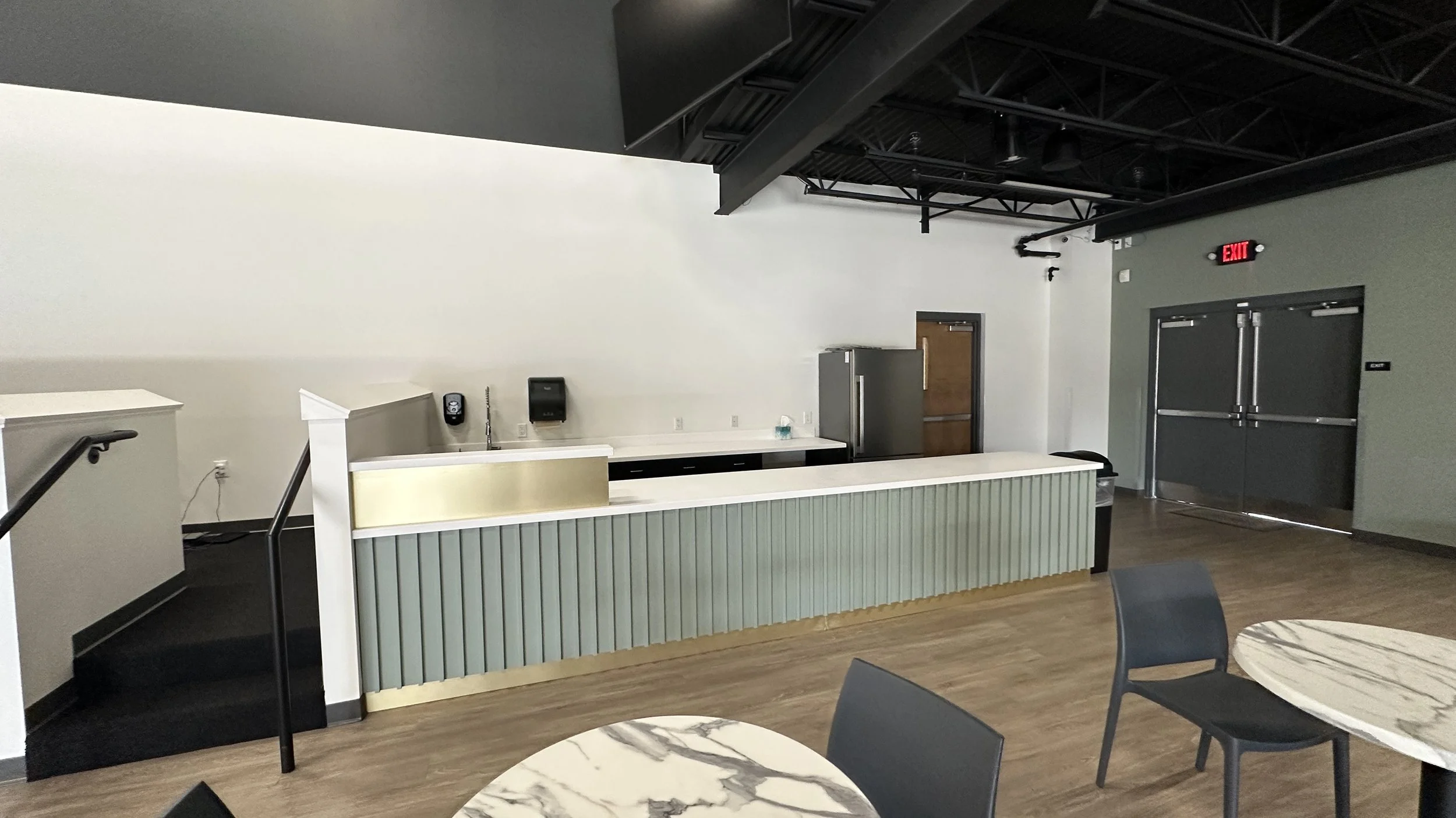 Empty cafeteria or lounge with a soft green counter, tables with marble tops, black chairs, and a plain white wall.