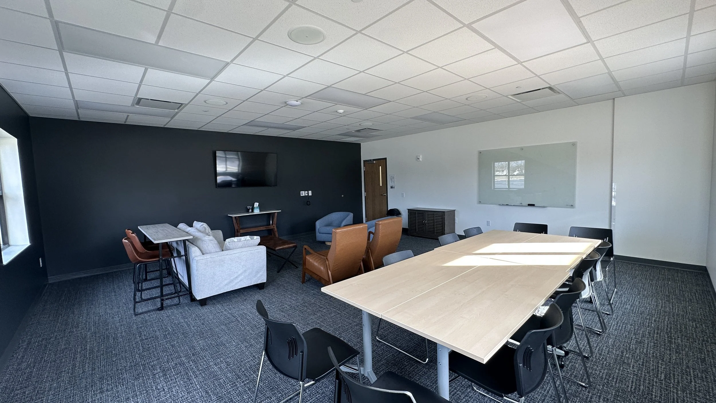 A modern conference room with a large wooden table surrounded by black chairs, a white couch and brown armchairs in a separate seating area, a wall-mounted TV, a whiteboard, and a window letting in natural light.