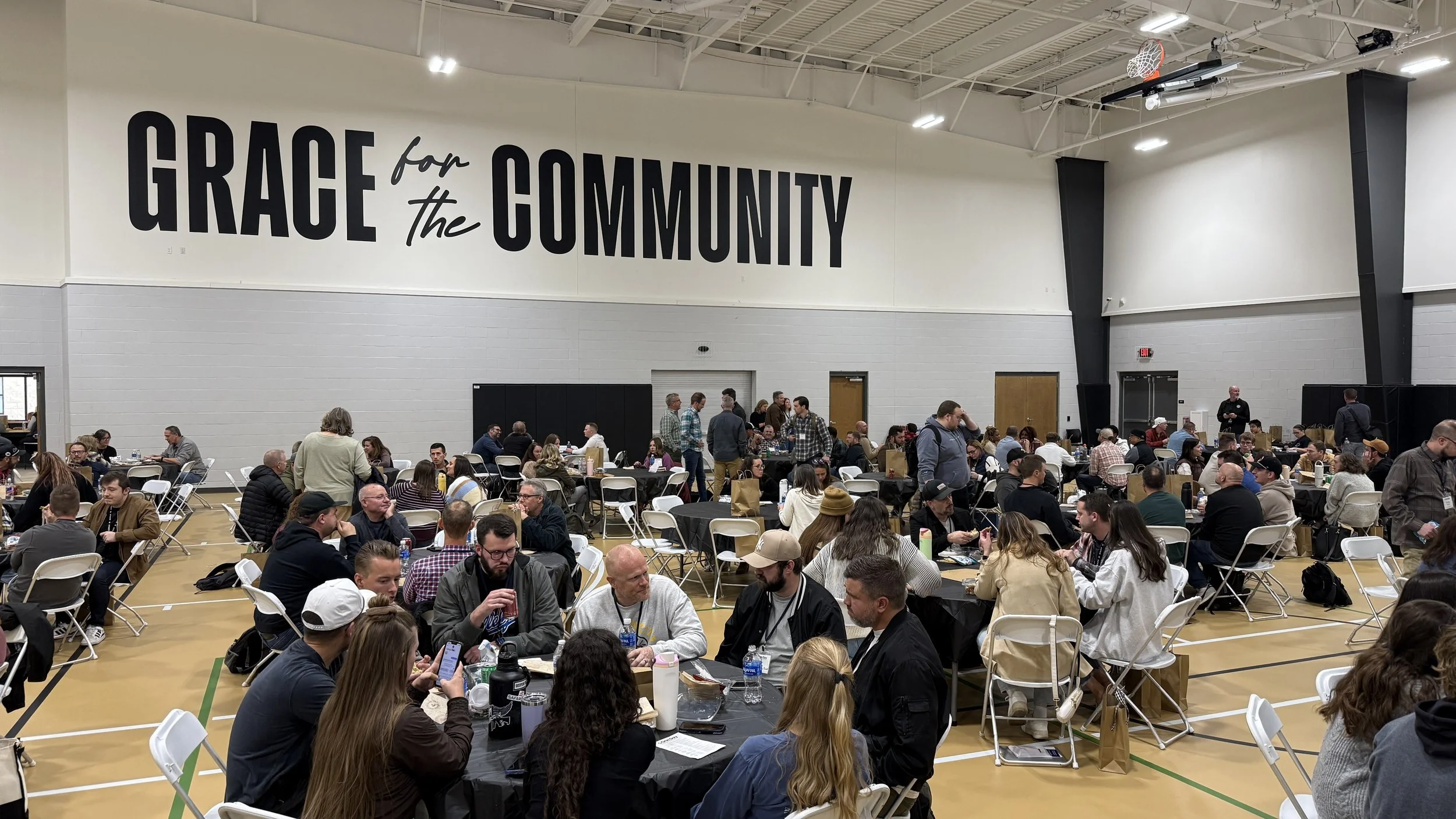 Large indoor gathering in a gymnasium with round tables and chairs, featuring many people seated, chatting, and engaging in activities. A prominent sign on the wall reads 'Grace for the Community'.