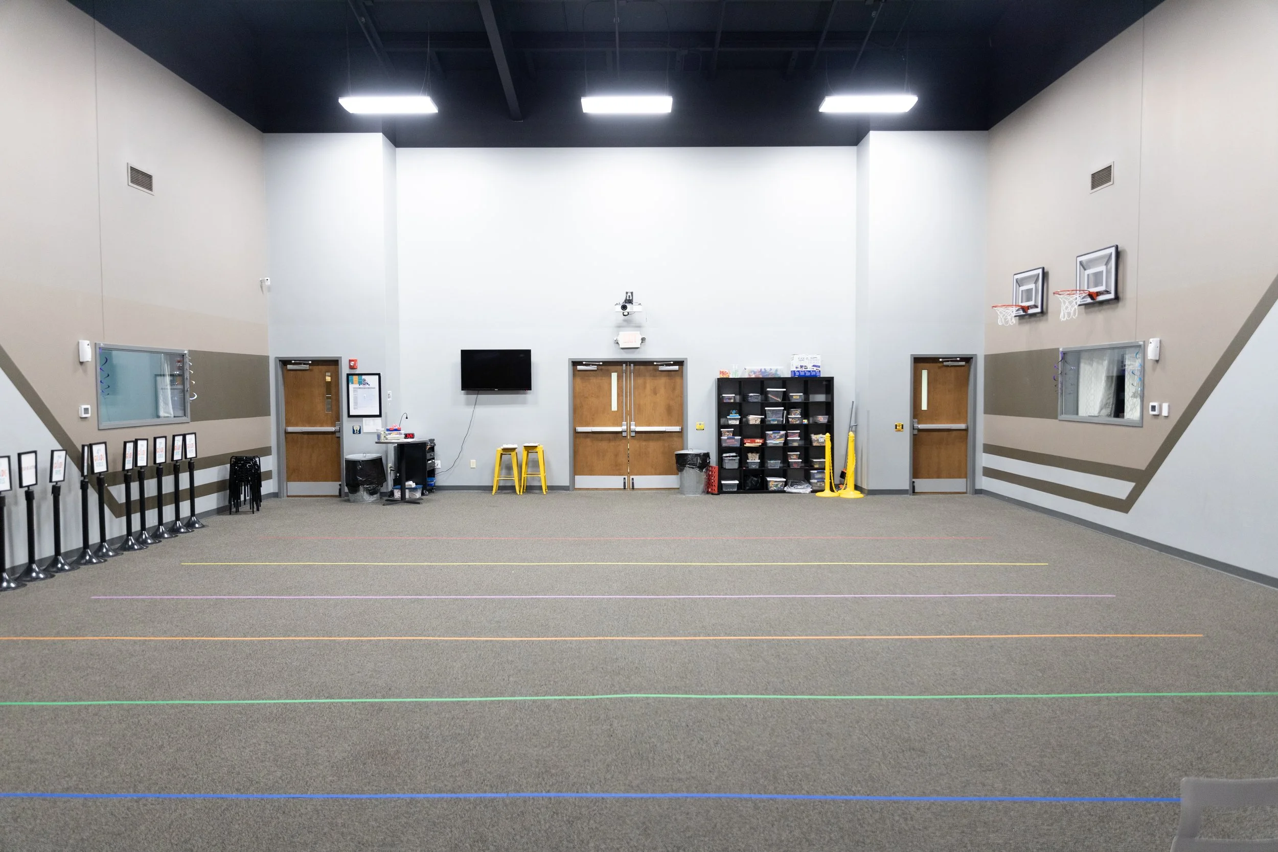 Indoor gymnasium with basketball hoops, a television screen, a shelving unit, and four doors, with colored lines on the carpeted floor for activities.