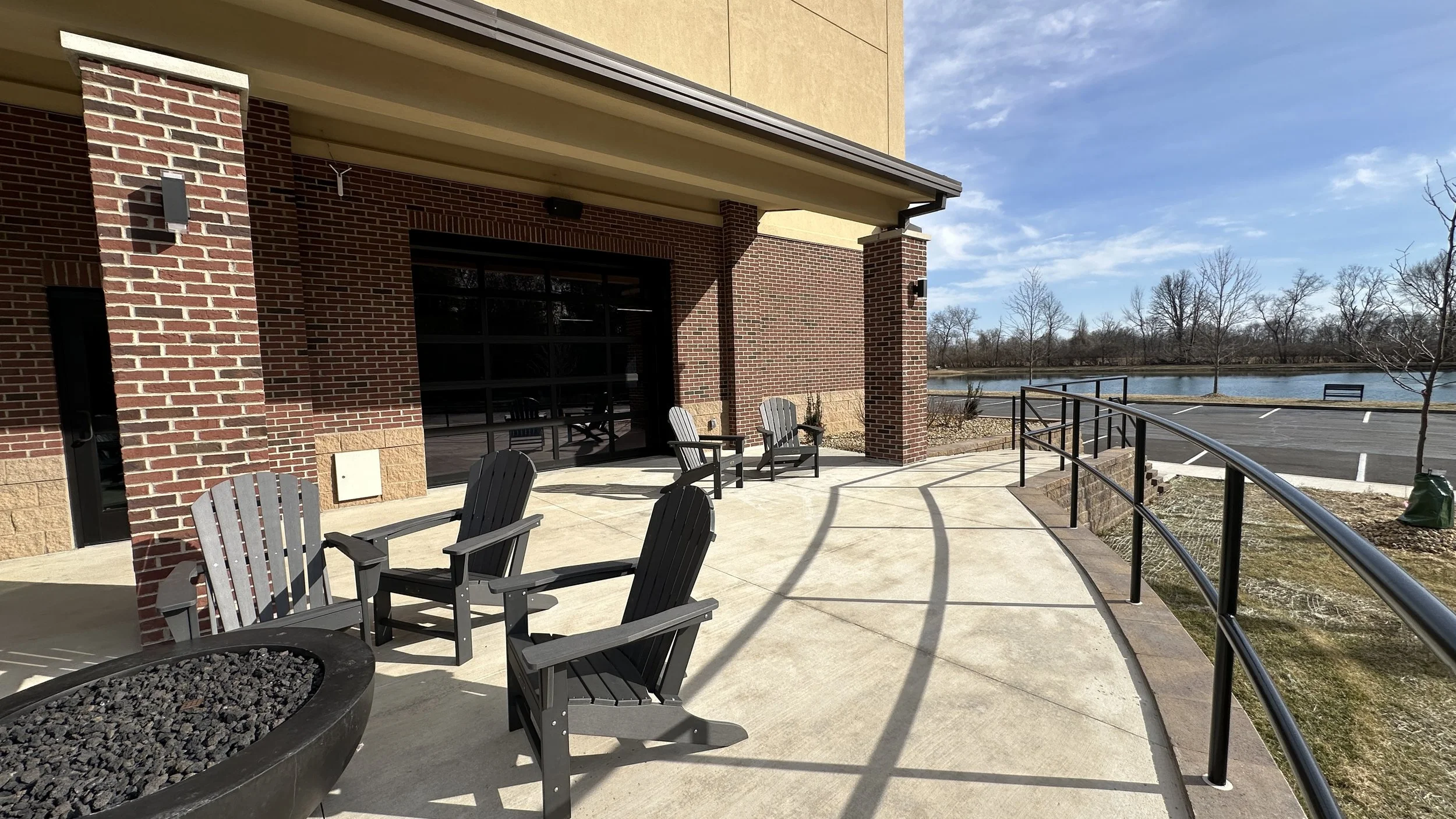 Outdoor patio with four black Adirondack chairs, a large concrete walkway, a metal railing, and a view of a pond with leafless trees in the background on a sunny day.