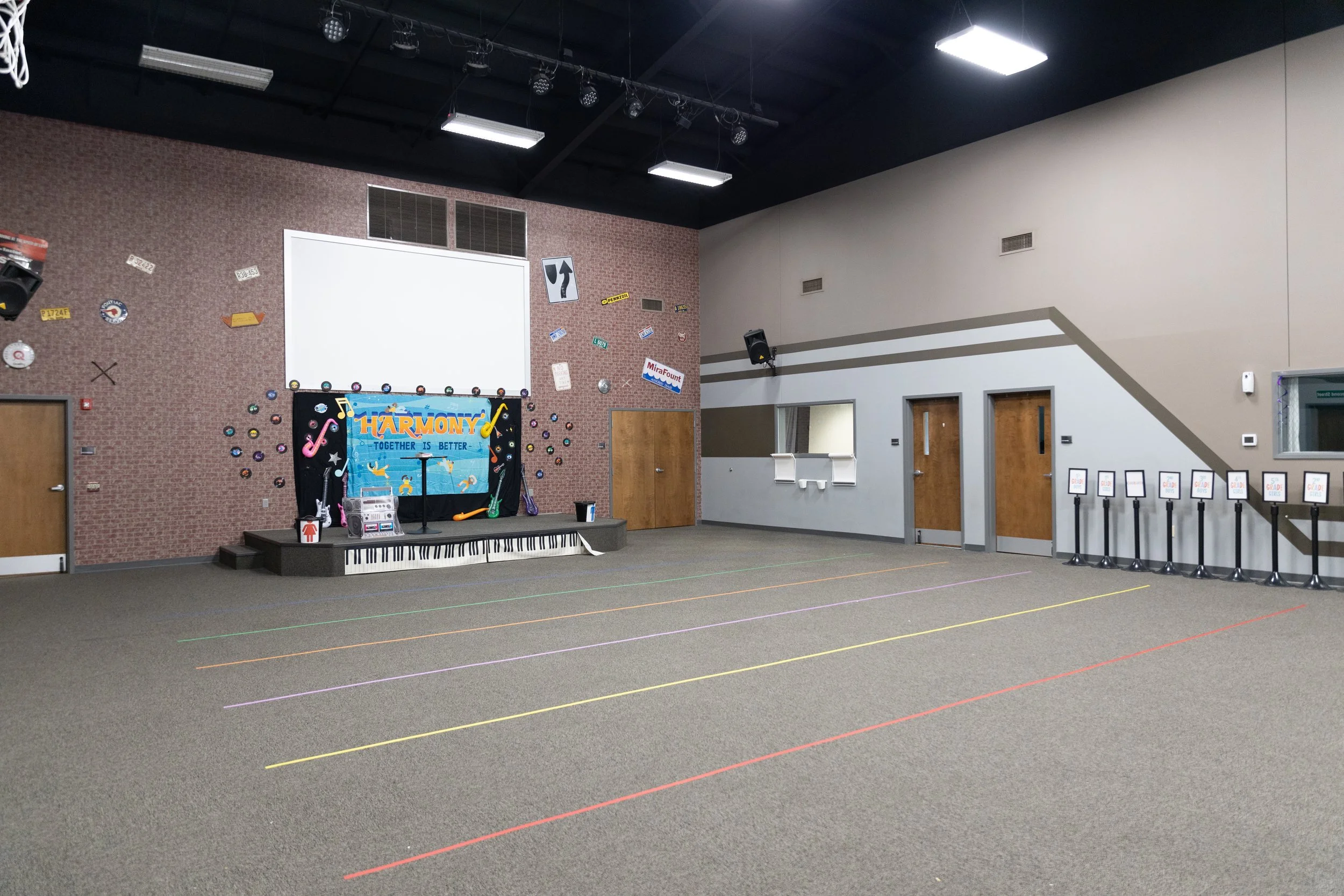 Empty indoor room with a stage decorated with a sign that says 'Harmony: Together is Better', musical notes, and colorful decorations. The room has a brick wall and a gray wall with doors, small windows, and a row of standees with signs. The floor is