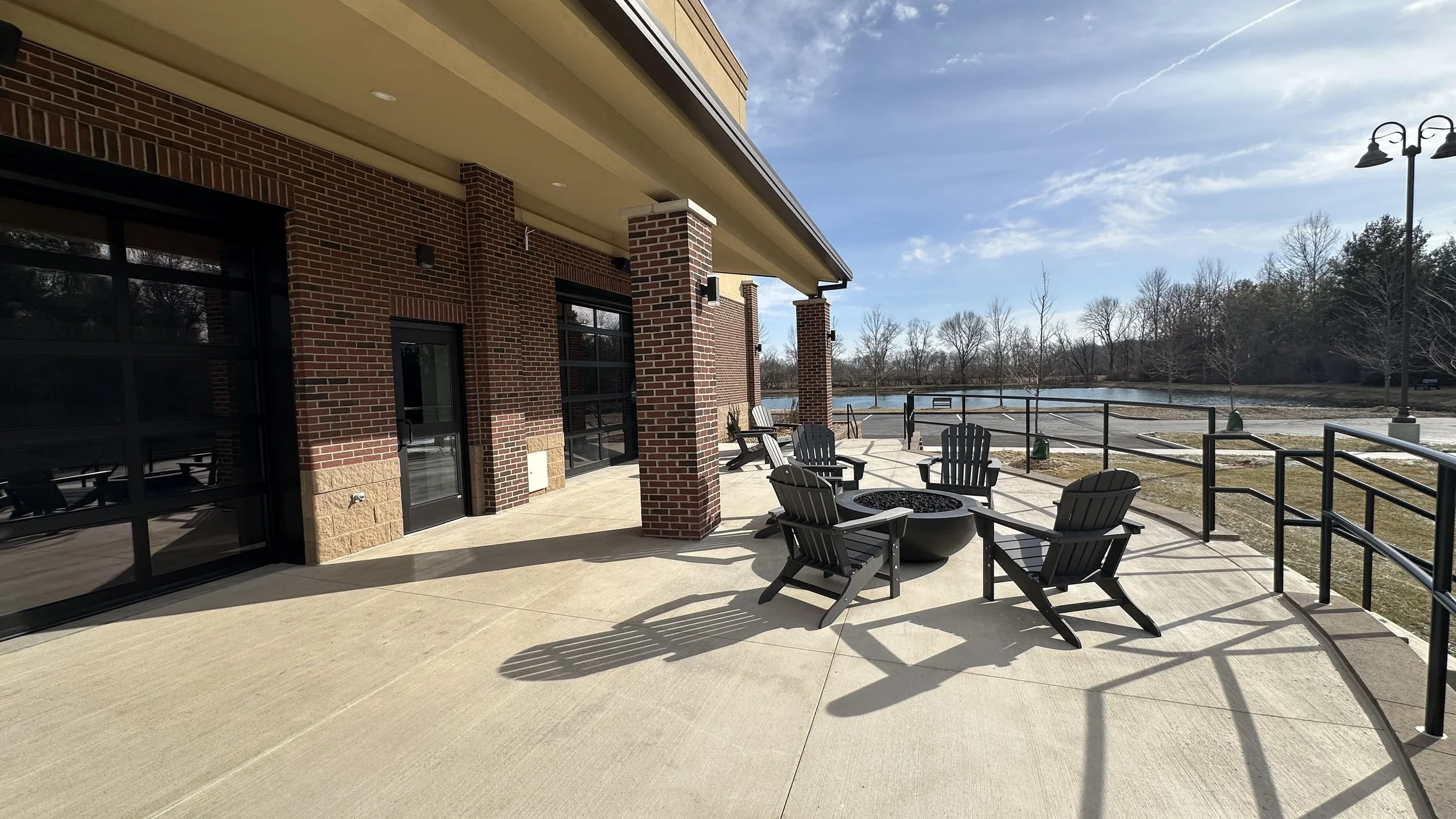 A spacious outdoor patio with black Adirondack chairs arranged around a fire pit, overlooking a lake with trees in the background on a sunny day.
