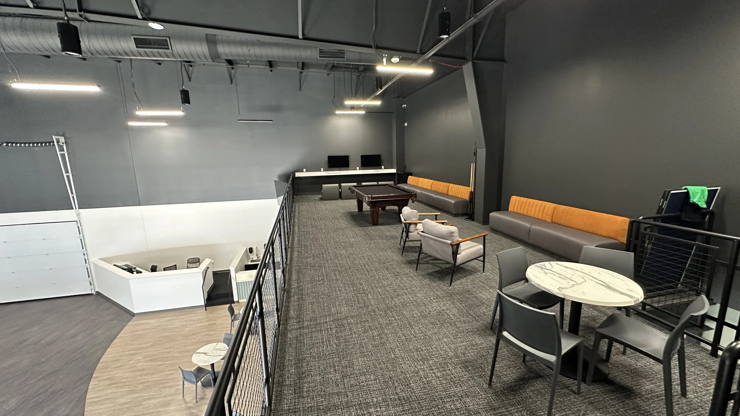 A spacious lounge area with dark gray walls and carpeted floor, featuring a pool table, multiple seating options including a long gray and orange banquette, armchairs, small round tables, and a gaming area with two monitors, with minimal lighting fro