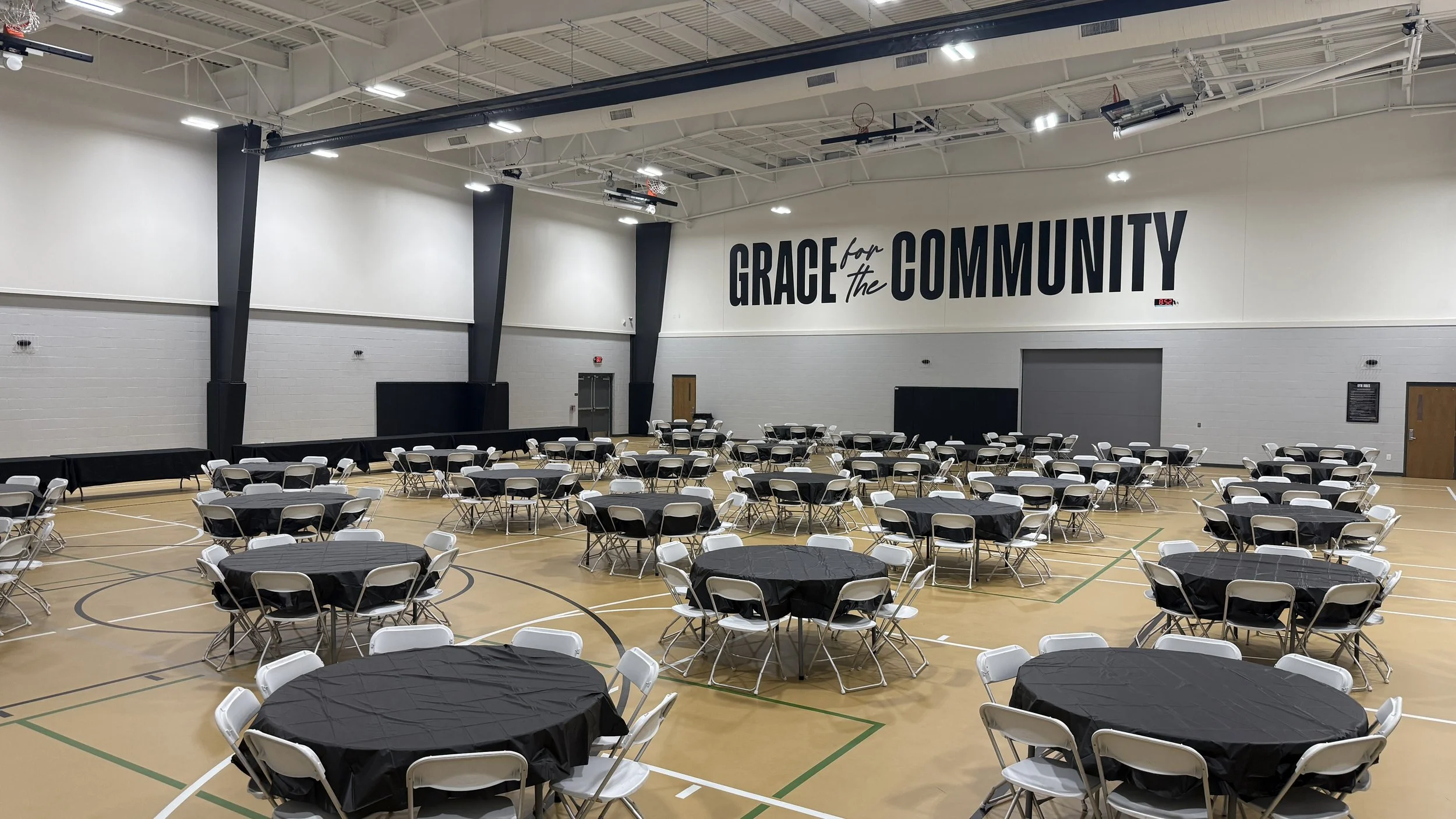An indoor gymnasium set up for an event with round tables covered with black tablecloths and white folding chairs, and a large wall with the phrase 'GRACE for the COMMUNITY' written on it.